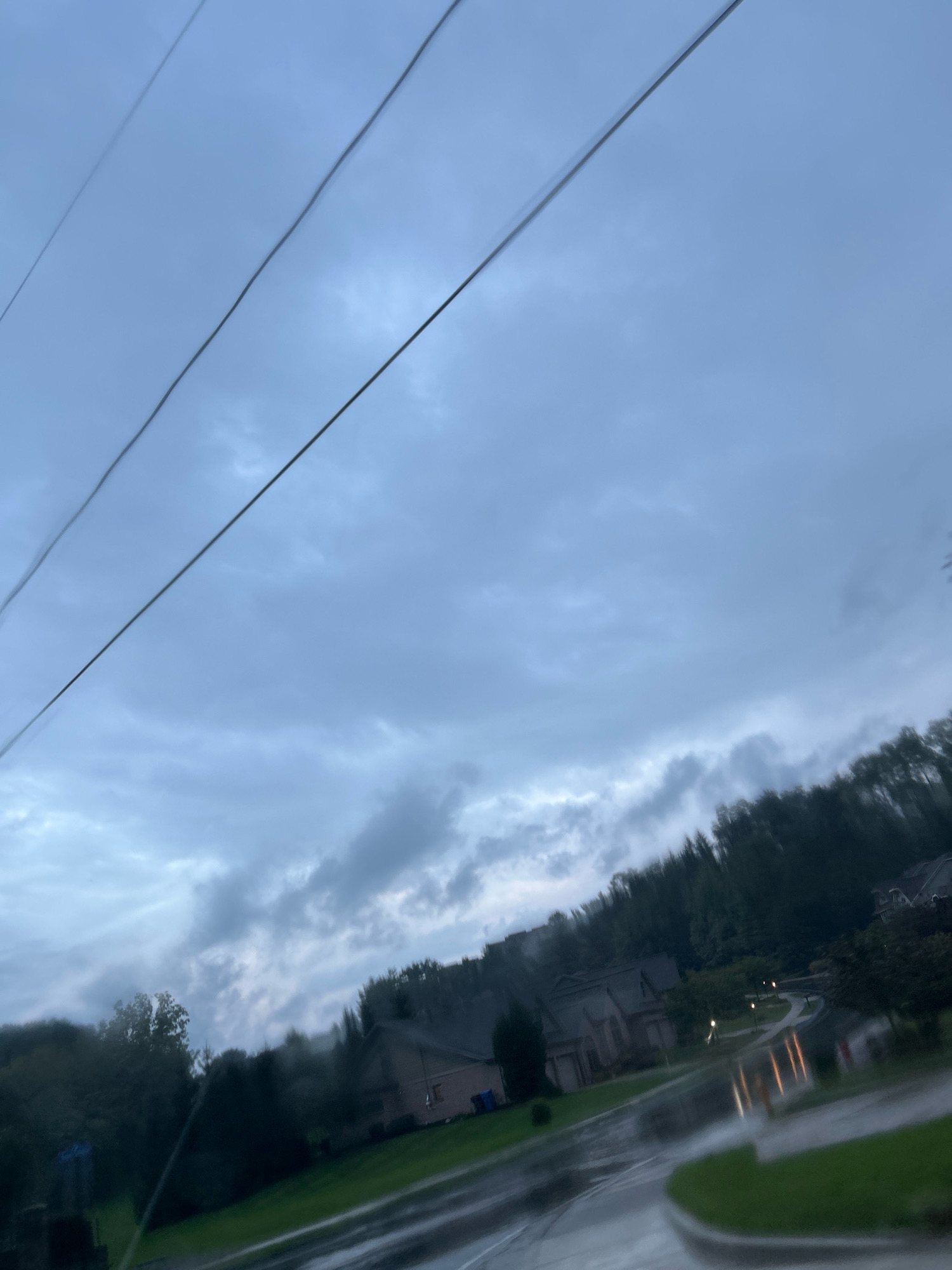 The image depicts a suburban street scene on an overcast day, likely in Pittsburgh, United States of America. Three parallel power lines stretch diagonally across the upper portion of the frame. Below, a residential neighborhood is visible with several houses situated on a hillside. The road and surrounding grass are wet, suggesting recent rainfall, with reflections of streetlights visible on the asphalt. Trees line the top of the hill, creating a dense backdrop. The overall atmosphere is muted and somewhat somber due to the cloudy sky and wet conditions.