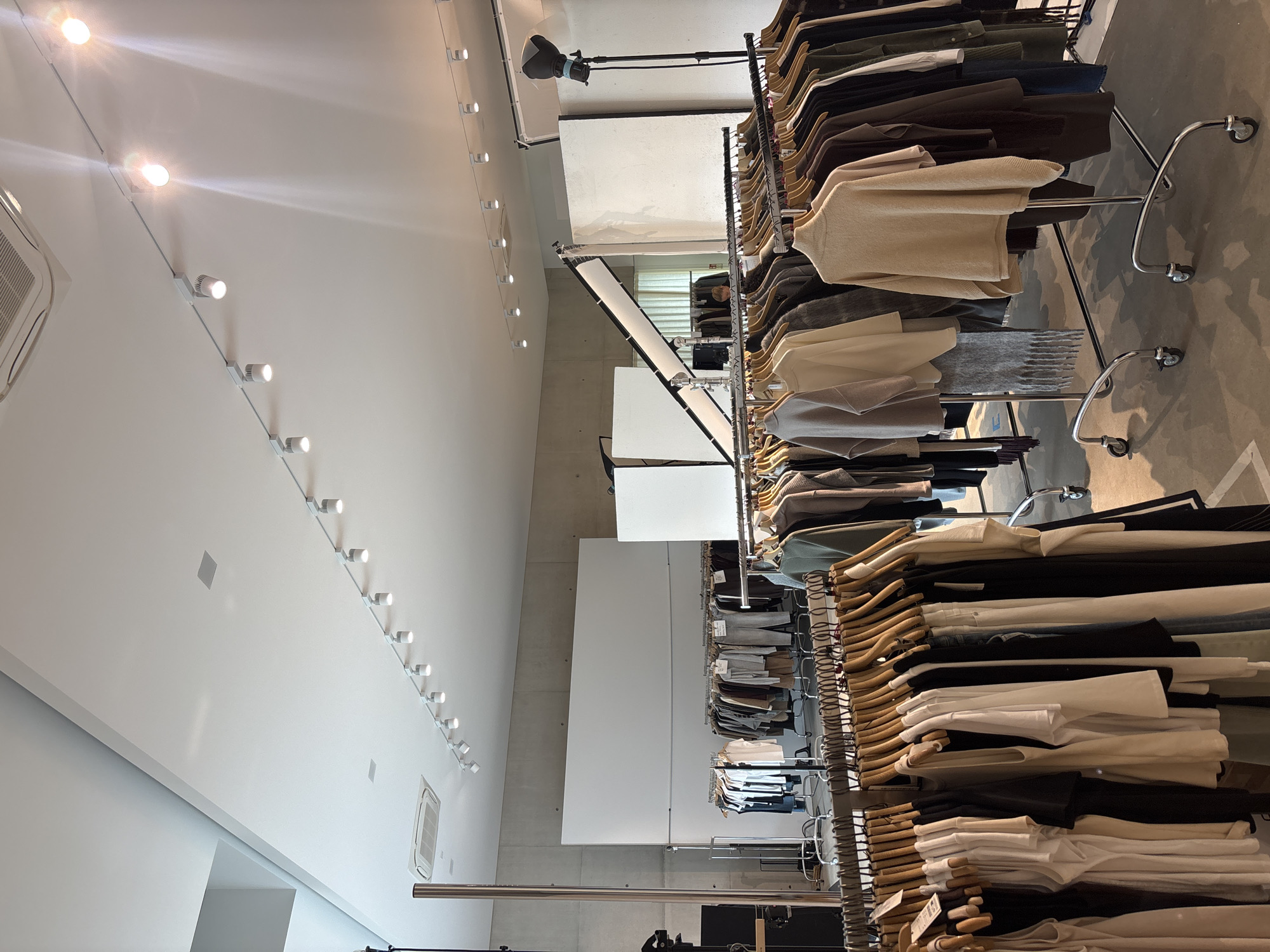 The image depicts the interior of a clothing store in Oelde, Germany. The scene is organized with racks of neatly hung apparel, primarily focusing on neutral-colored garments such as sweaters, shirts, and pants. The lighting in the store is a mix of overhead track lighting and accent lights, creating a bright and airy atmosphere. The floor appears to be concrete, a common feature in modern retail spaces.
Multiple racks of clothing are visible, suggesting a wide selection of items available for purchase. The clothing is predominantly in shades of beige, cream, white, gray, and black, giving the store a minimalist and sophisticated aesthetic. Some garments are folded on shelves within the racks, while others are displayed on hangers. A few accessories, like a grey fringed scarf, are also interspersed among the clothing.
The activity taking place is the display and organization of clothing for sale. There are no people prominently featured in the image, indicating the photograph might have been taken before or after operating hours, or focusing solely on the merchandise and store design. The setting is clean and well-maintained, suggesting a professional retail environment.
Notable details include the track lighting system running along the ceiling, various light fixtures, and an air conditioning unit mounted high on the wall. The metal racks on wheels suggest a flexible store layout. The overall impression is one of a contemporary boutique offering fashionable, understated clothing. No visible text is present in the image.