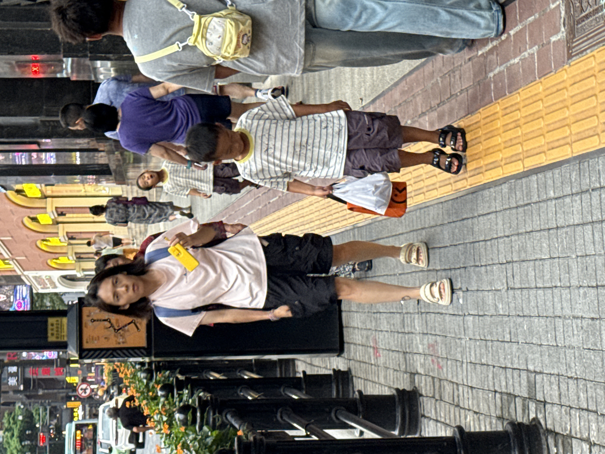 A woman and a young boy walk along a city sidewalk in Yuzhong District, China. The woman, wearing a light pink t-shirt, black cargo shorts, and sandals, carries a yellow object in her right hand and a blue backpack. The boy, dressed in a striped t-shirt and dark shorts with sandals, holds a white and orange shopping bag in his left hand. They are walking on a paved sidewalk with a tactile paving strip. In the background, other pedestrians are visible, along with storefronts and street signs. The scene depicts an urban environment with shops and people going about their day.