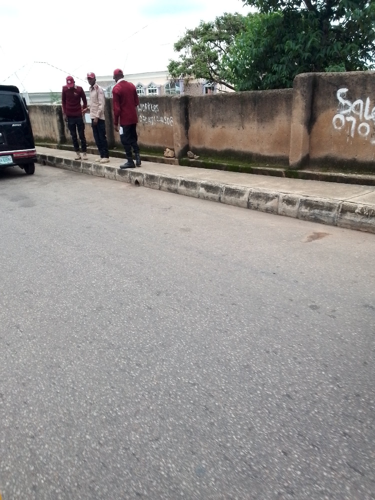 This image depicts a street scene in Jos, Nigeria. In the foreground, a gray asphalt road with scattered white aggregate is visible. A concrete sidewalk runs along the right side of the road, bordered by a low concrete curb. Behind the curb, a weathered brown concrete wall runs horizontally across the image. Graffiti, including the word "WORKERS" and a phone number, is spray-painted on the wall. To the left of the wall, the upper portion of a white building with arched windows can be seen. Lush green trees with dense foliage are present above and behind the wall.
On the sidewalk, three individuals are standing. The person on the far left wears a dark red shirt and dark pants, with a red cap. Next to them, a person in a light tan shirt and dark pants, also wearing a red cap, stands facing away from the camera. The person on the far right is dressed in a dark red jacket and black pants, wearing a red cap and black shoes. They appear to be engaged in a conversation. A black vehicle is parked on the left side of the road, facing away from the viewer, with its rear visible. A license plate, partially obscured, reads "YKK 506NC". The sky is overcast and light gray.