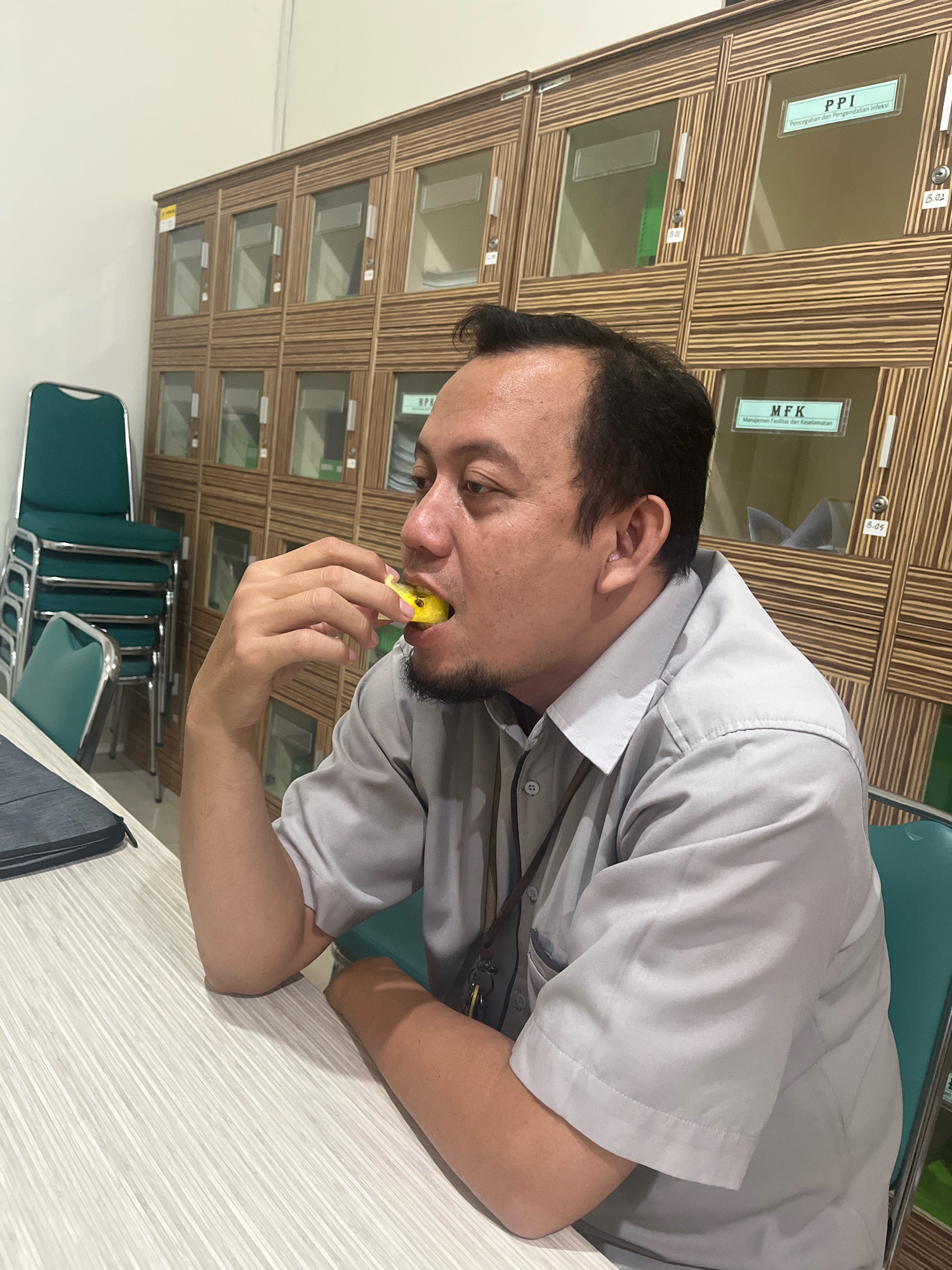 A man is sitting at a table with a laptop on it, eating a yellow object. He is wearing a light gray collared shirt. Behind him is a wall of wooden lockers with glass doors, some of which have labels. Stacked green chairs are visible to the left of the lockers. The scene appears to be indoors, possibly in an office or storage area. The location is Dapur, Indonesia.