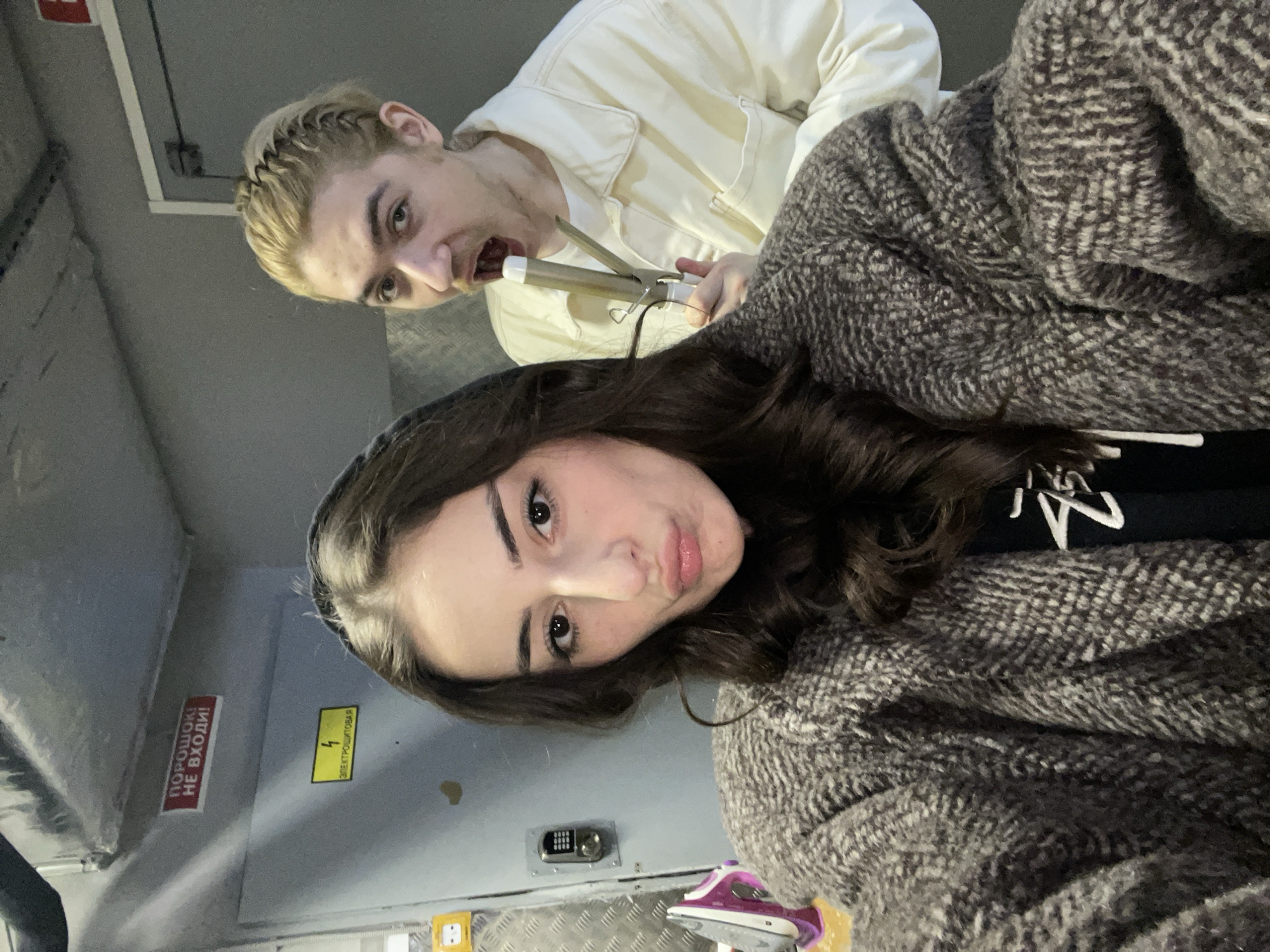 The image depicts two people posing in what appears to be an industrial or utility area, possibly the inside of a building or compartment. The person in the foreground is a young woman with dark, wavy hair, dark eyebrows, and a light complexion. She is wearing a dark-colored shirt with a white design and a large, textured gray and white patterned coat or sweater. She is looking directly at the camera with a slight pout.
Behind her is a young man with blond hair braided at the front, wearing a white jacket. He is holding a curling iron in his mouth and hand, striking a silly pose with his eyes wide and mouth open.
The background is primarily metallic gray and includes warning signs, a numerical keypad, and electrical outlets. The signs are in Russian, with one appearing to say "ПОРОШОК! НЕ ВХОДИ!" (POWDER! DO NOT ENTER!) and another with a lightning bolt symbol denoting electrical hazards. The environment suggests an industrial or utility setting in a Russian-speaking area, possibly Moscow.