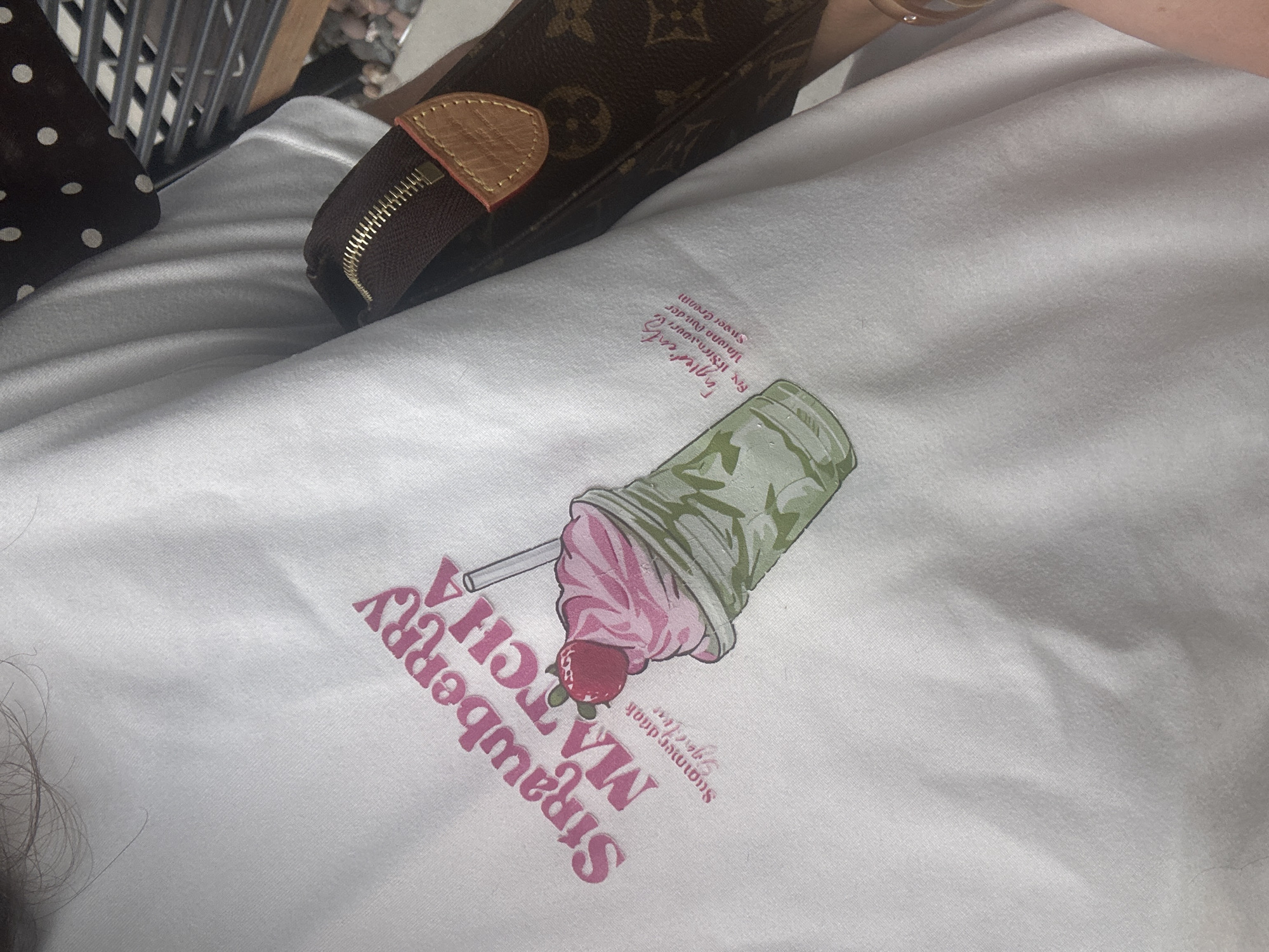 The image shows a close-up of a white t-shirt featuring a graphic of a strawberry matcha drink. The graphic includes an image of the drink with a straw, strawberry, and associated text. A brown wallet and polka dot item are also partially visible in the background.