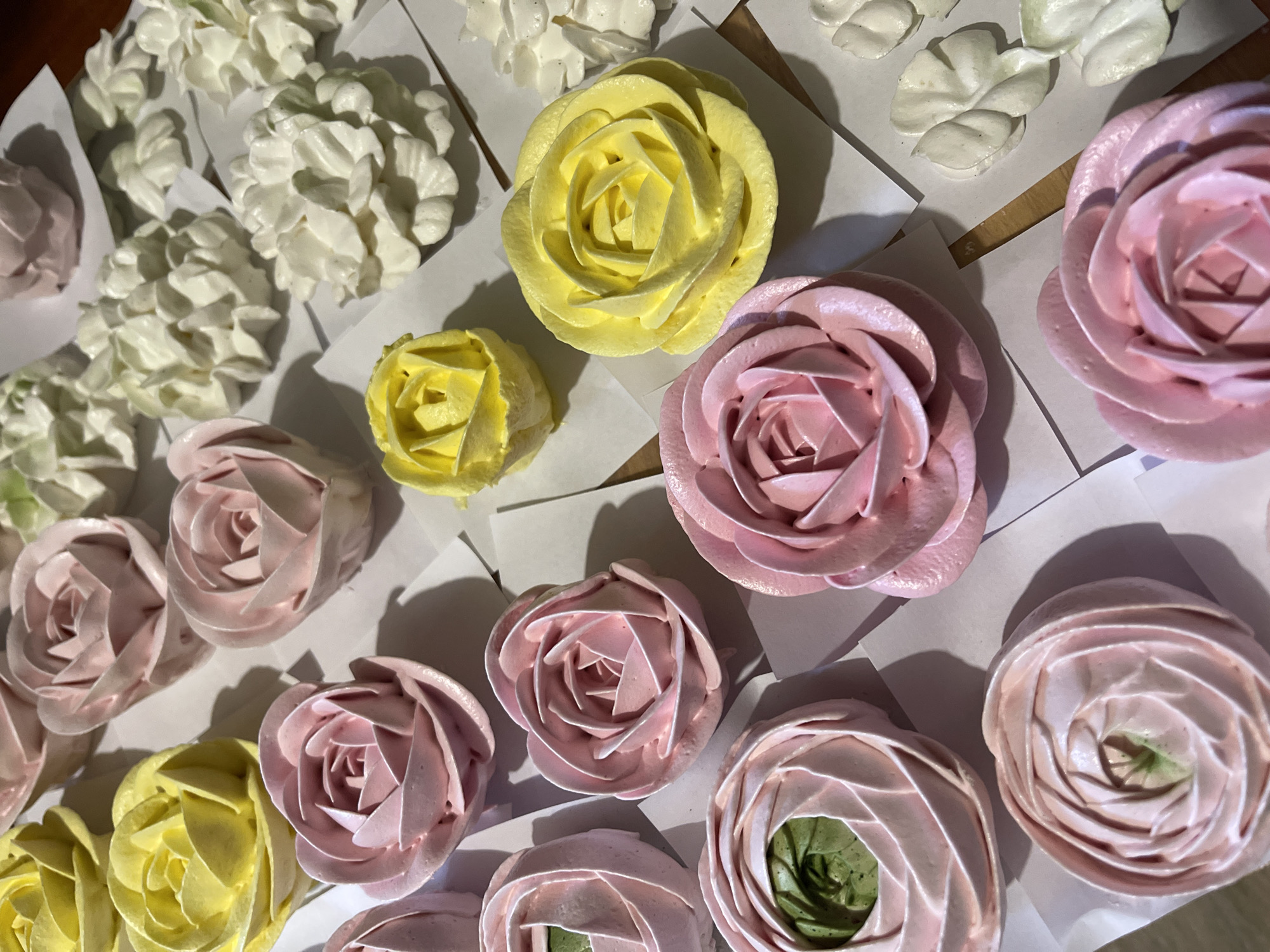 A close-up, overhead shot shows an assortment of meringue cookies piped into the shape of roses and other flower-like designs. The meringues are arranged on white paper squares, some of which are placed on a wooden surface. The colors of the meringues include pale yellow, various shades of pink, and white. Some of the pink meringues have a hint of green in their center, suggesting a specific color variation or shading. The lighting is somewhat dim, creating shadows that add depth to the image. The perspective is straight down, focusing on the texture and intricate piping of the meringue creations.