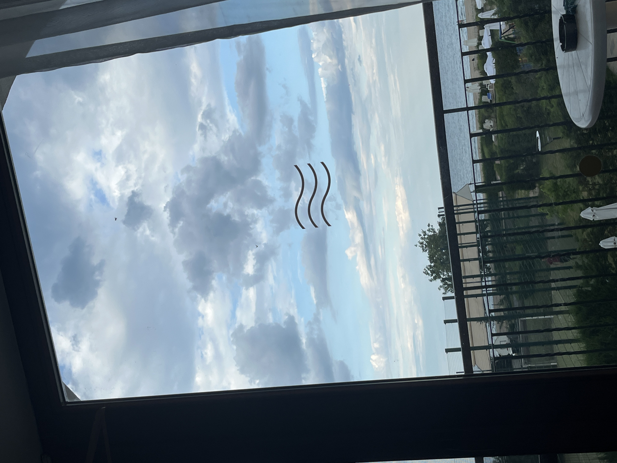 The image, taken from inside a room looking out through a window or balcony, shows a view of the Black Sea coast in Obzor, Bulgaria. The sky is cloudy with patches of blue, and there are three horizontal wavy lines superimposed on the sky, possibly a graphic element. Below the sky is the sea, which appears calm. In the middle ground, there is a railing, a green fence, trees, and what appears to be a beach with white umbrellas and lounge chairs. A white table with an ashtray is visible in the foreground. The scene depicts a cloudy day at a seaside resort.