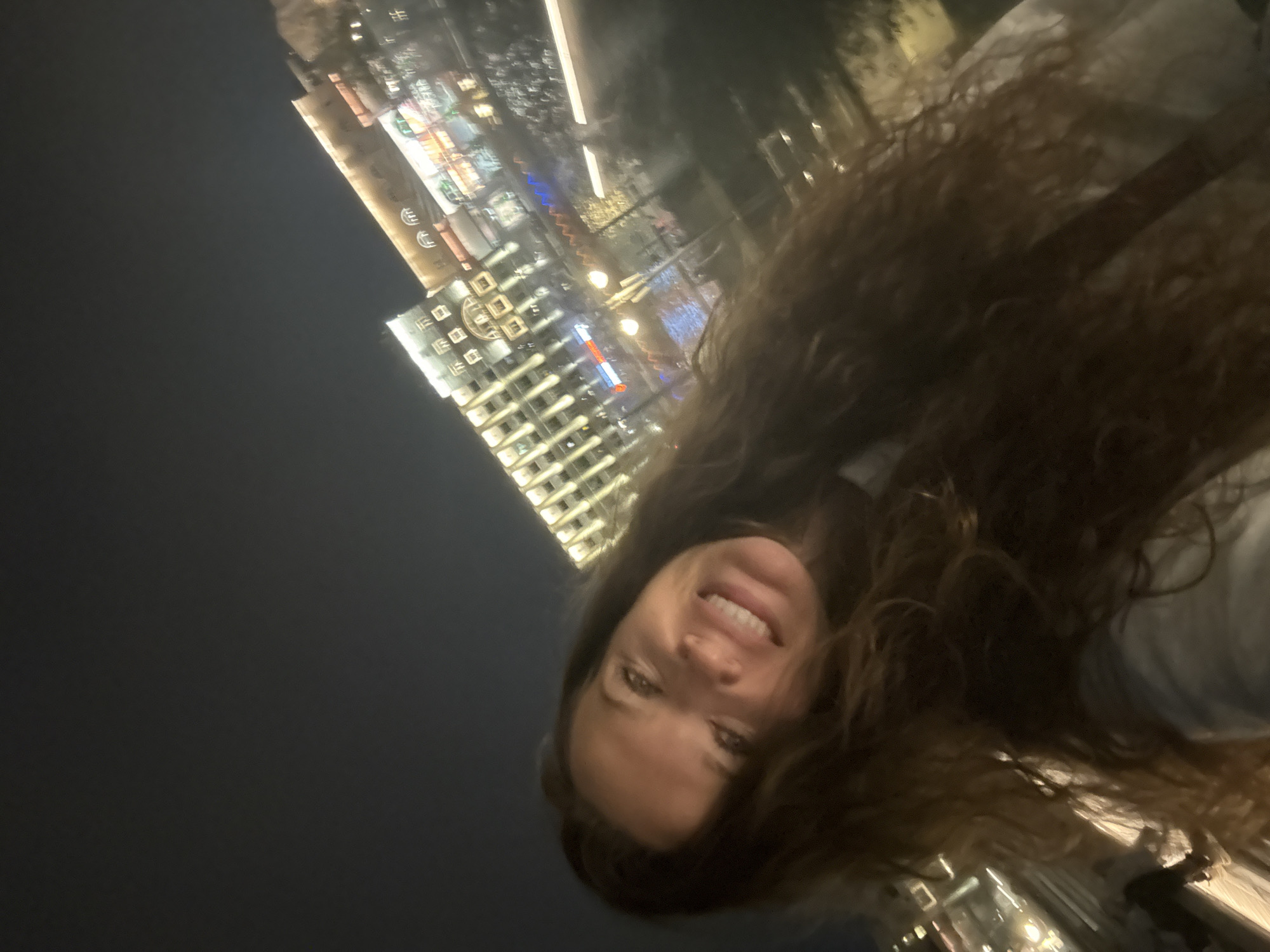 A woman is taking a selfie at night with a city skyline in the background. The woman has long, dark, wavy hair and is smiling at the camera. She is wearing a light-colored top. In the background, there is a large, multi-story building illuminated with many lights. The building appears to be a hotel or a commercial establishment with numerous windows and architectural details. There is also a body of water, possibly a river or canal, reflecting the city lights. The overall scene suggests a vibrant urban environment at night. It is likely located near the Moscow River, with the illuminated building possibly being a hotel or entertainment complex along the embankment.