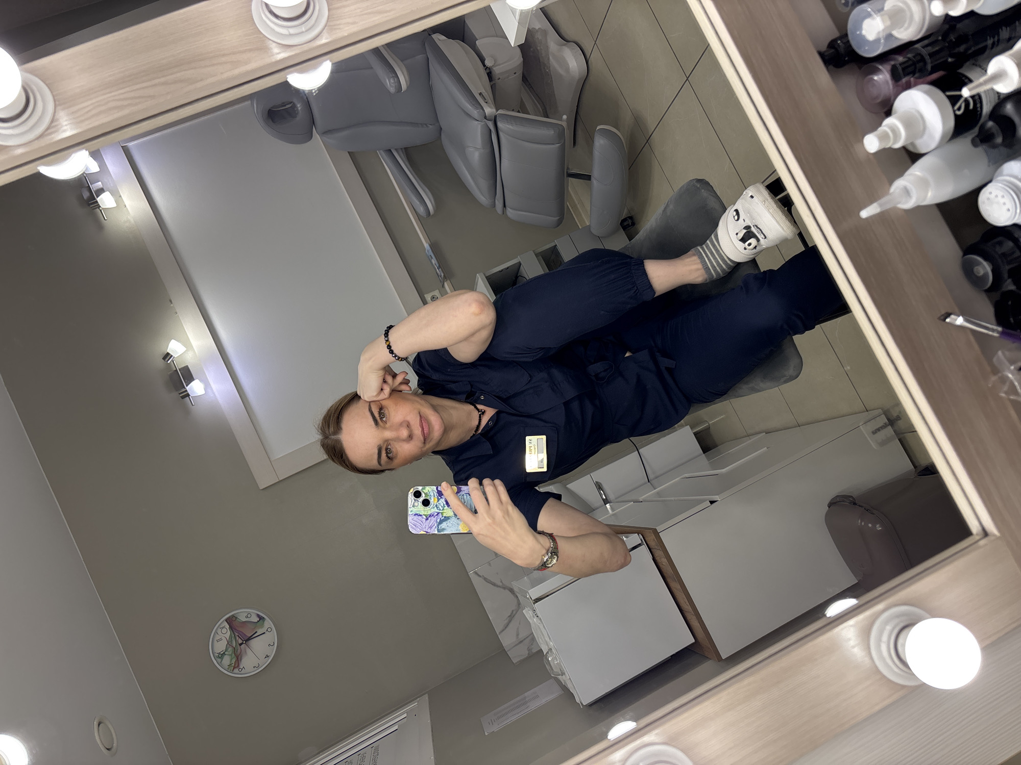 A mirror selfie of a woman in a dark blue jumpsuit, seated in a room with a dental chair, is captured. The woman, who is fair-skinned, holds a patterned phone up to take the picture. The room includes a clock on the wall, a white sink, and bottles of chemicals. Her attire includes a name tag and a watch. One of her legs is visible with a cartoon print sock. The room is likely a dental clinic based on the presence of the chair. The mirror has lights attached around its perimeter.