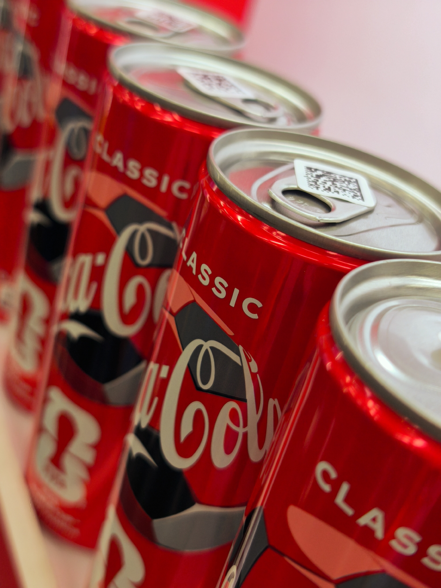 The image shows a close-up, shallow depth-of-field, angled view of multiple Coca-Cola cans arranged in a row. The cans are predominantly red with the classic white Coca-Cola logo and the word "CLASSIC" printed on them. Some cans have a black and gray design element. The tops of the cans are visible, showing the silver pull tabs and some small white stickers with QR codes. The background is a soft, blurred pinkish hue. The scene does not depict any identifiable urban or geographical features of Tashkent, Uzbekistan. There are no people, actions, or interactions shown. The focus is entirely on the product.