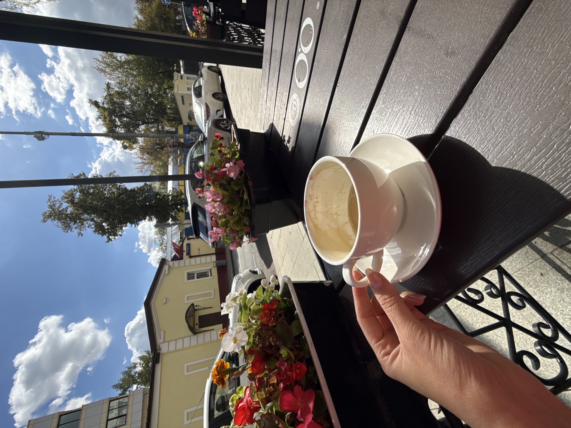 An outdoor cafe table in Moscow, Russia, is set with a white ceramic cup of coffee and saucer. A woman's hand, with manicured nails, is seen gently holding the cup. In the foreground, a planter box overflows with red and pink flowers, casting shadows on the dark wooden table. Beyond the table, a street scene unfolds with parked cars and buildings under a bright blue sky dotted with fluffy white clouds. The architecture suggests a blend of older, low-rise yellow buildings and a more modern, taller structure. The overall atmosphere is one of a pleasant, sunny day spent enjoying a coffee break.