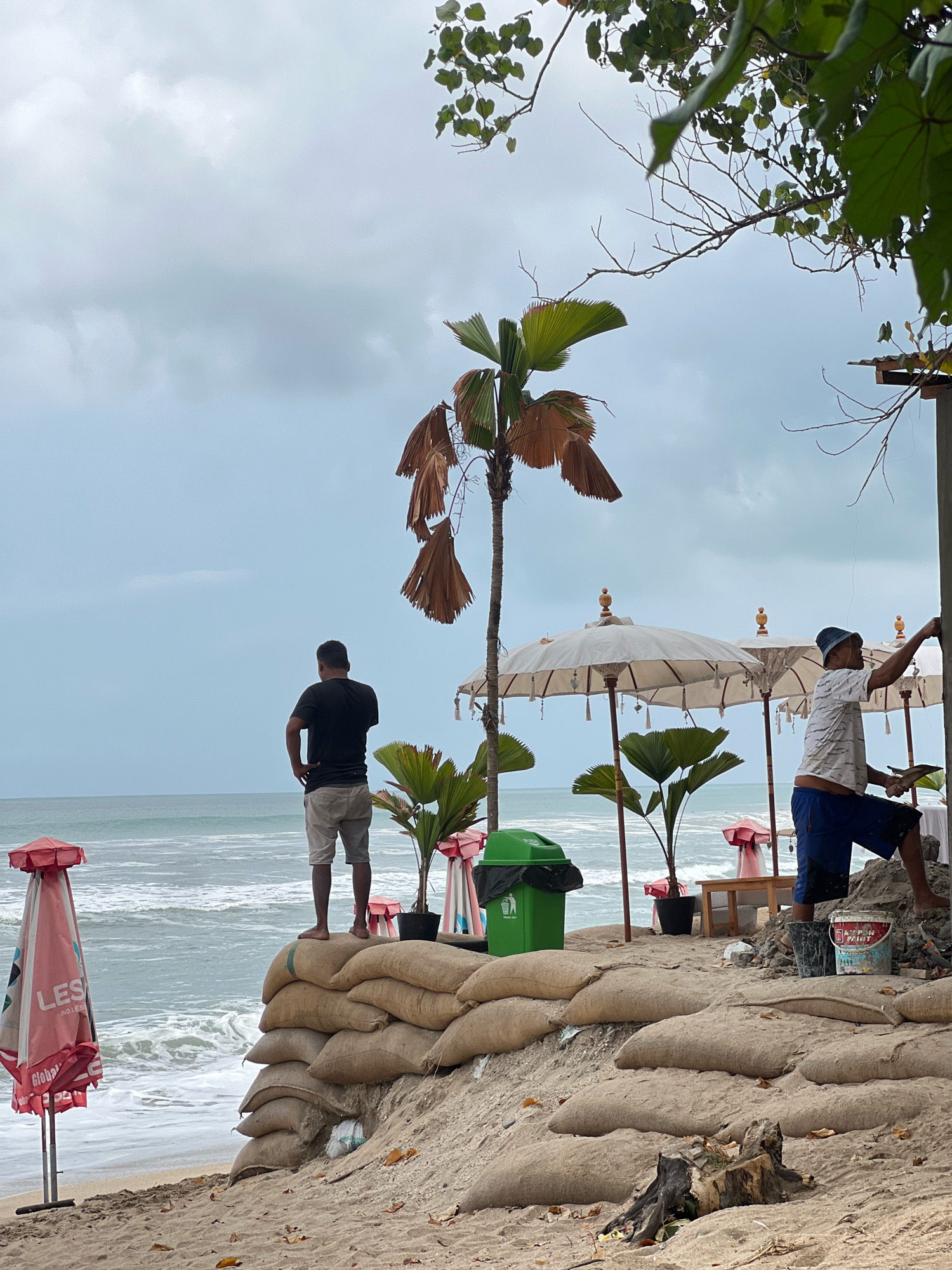 The image depicts a beach scene in Kuta, Indonesia, on an overcast day. In the foreground, sandbags are stacked in a berm, likely as a measure against coastal erosion. A partially folded red umbrella with "LES" visible on it stands to the left. Further back, two men are present. One man, wearing a black t-shirt and shorts, stands with his back to the camera, looking out at the ocean from atop the sandbag berm. Another man, wearing a patterned shirt and blue shorts, is working near a structure, possibly painting or plastering a pole. White beach umbrellas are set up behind him. Palm trees, some with dried fronds, and green potted plants are scattered around the area. A green trash bin is visible next to the sandbags. The ocean waves are visible in the background, meeting the sandy shore. The sky is cloudy and muted in color.