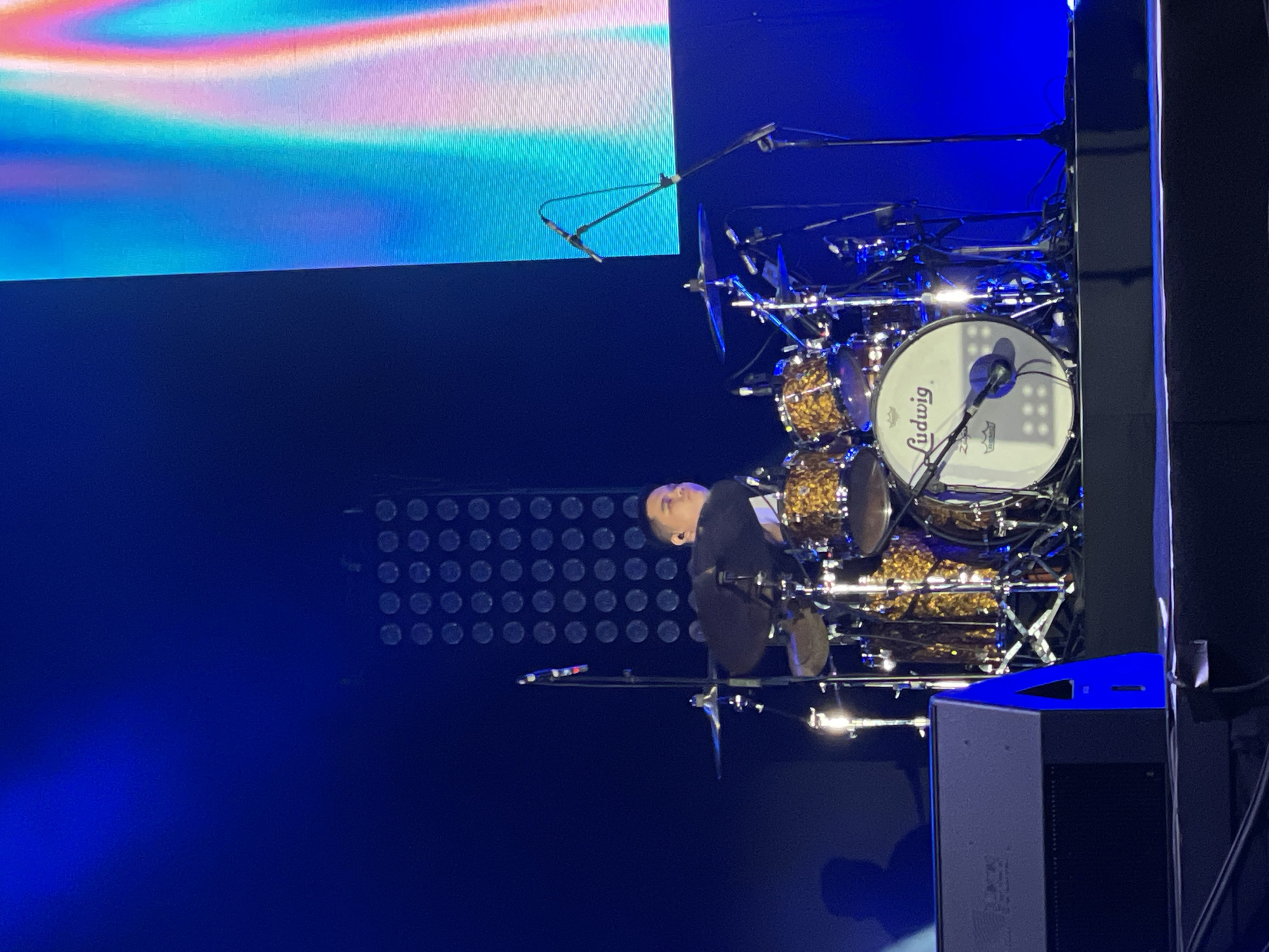The image shows a drummer performing on stage. He is seated behind a gold-colored Ludwig drum set, wearing a dark suit. The drums are fully equipped with cymbals and microphones. The backdrop includes a large LED screen displaying abstract colorful visuals and a lighting rig with circular light patterns. Speakers are positioned at the right edge of the frame. The location is Salembaran, Indonesia.