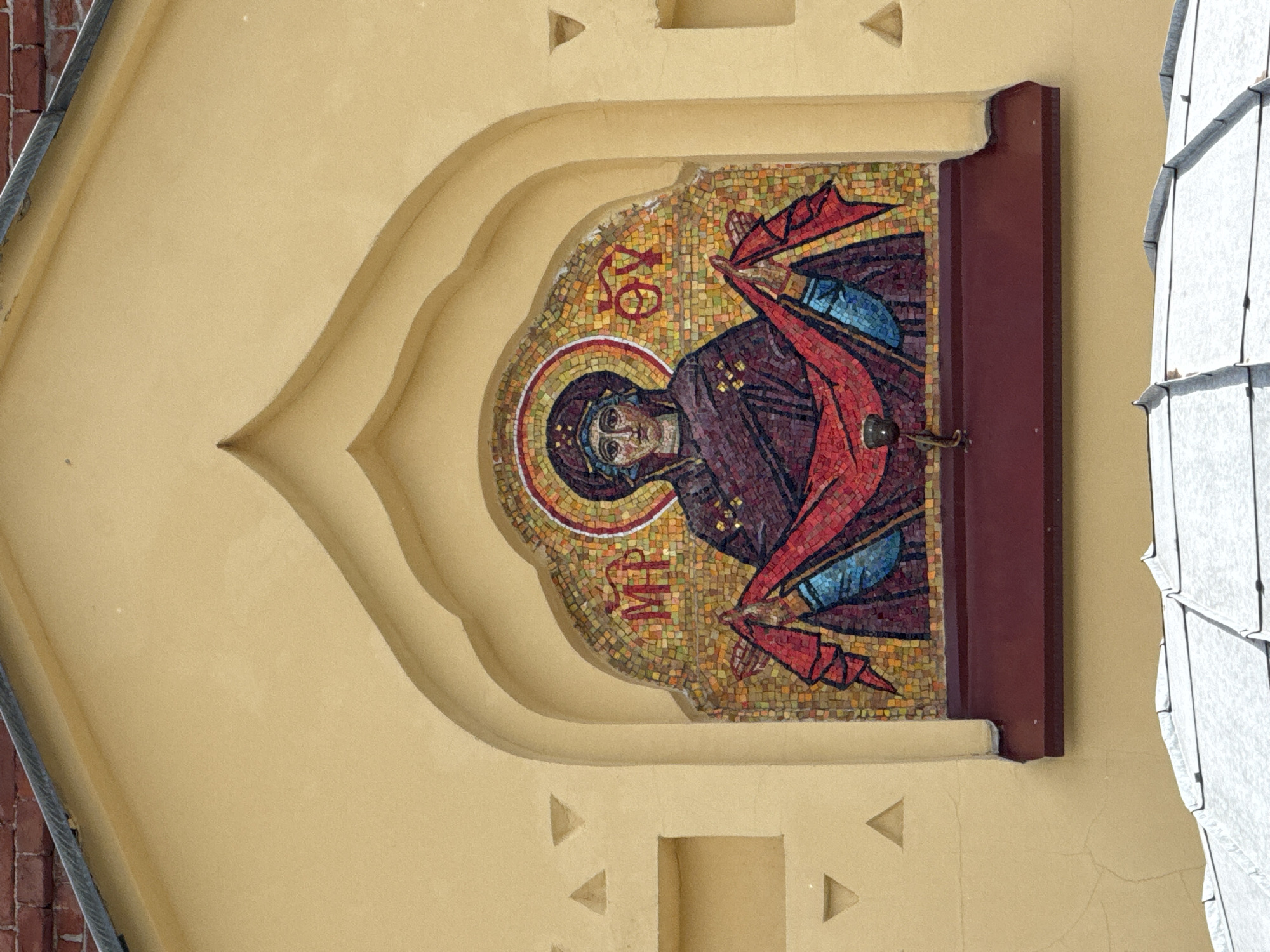 This is a photograph of a mosaic icon of the Virgin Mary, mounted on a yellow stucco wall. The mosaic is framed by a reddish-brown border and is set within a decorative arched niche. The icon depicts the Virgin Mary in traditional Byzantine style, with a halo surrounding her head. She is wearing a dark purple robe and a blue veil. Her hands are raised, and she is holding a red cloth. Greek letters are visible within the halo and above her head, which are commonly found in Orthodox icons.
The surrounding wall is a light yellow color, and the architectural style of the building suggests it is likely a church or religious institution. The niche has a decorative, layered effect. To the left of the mosaic, a portion of a red brick wall is visible, indicating the building's construction. To the right, a white, textured surface, possibly roofing tiles or architectural cladding, is visible.
The lighting suggests it is daytime, and the shadows are not particularly harsh, indicating it might be overcast or the sun is not directly overhead. The overall impression is one of religious devotion and artistry. The photo is taken from a slightly low angle, looking up at the mosaic. The presence of the brickwork and the architectural style are consistent with a location in Saint Petersburg, Russia.