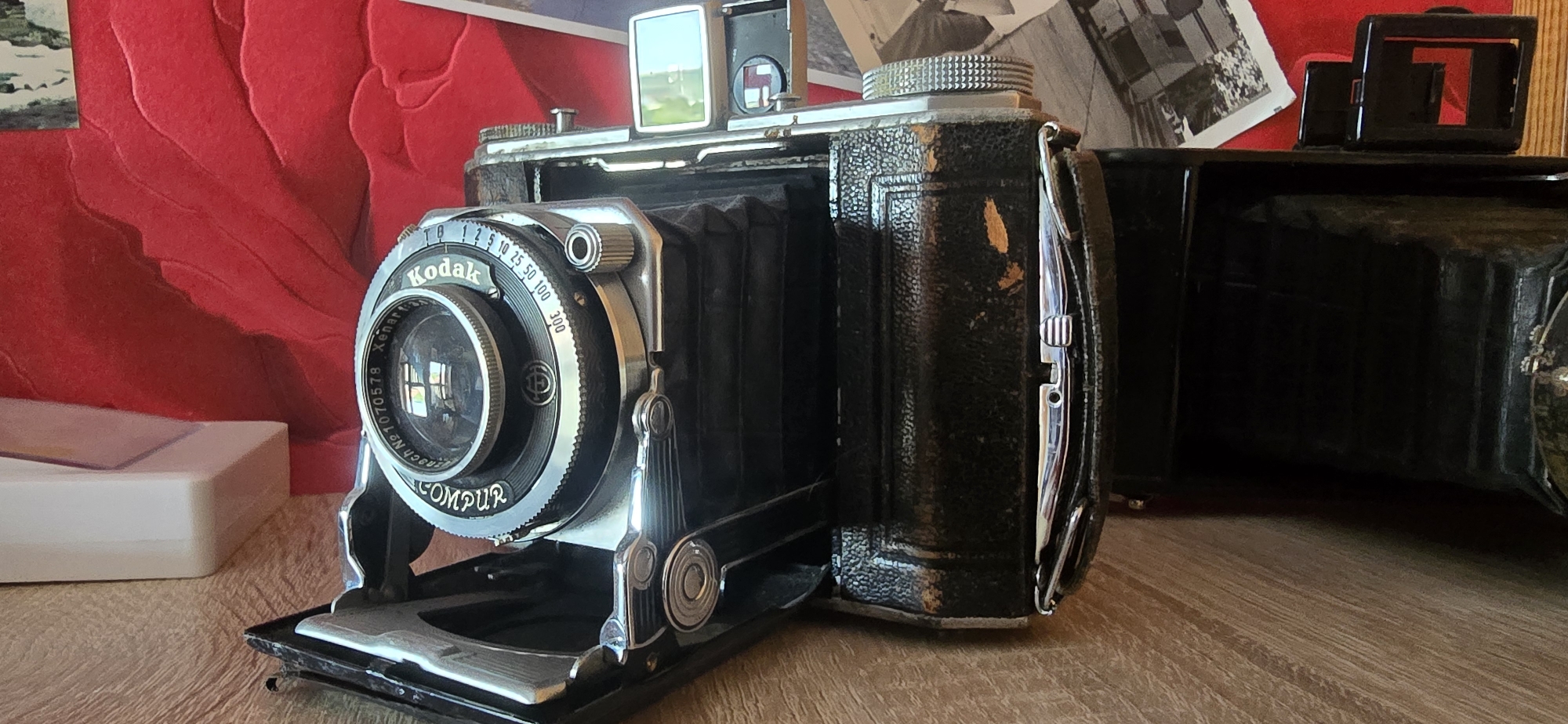 The image shows a vintage Kodak camera with a bellows, placed on a wooden surface. The camera is black with silver accents and has the brand name "Kodak" and "COMPUR" visible on the lens assembly. A red, textured background is behind the camera, with some black and white photographs or prints visible. A white object, possibly a camera accessory or case, is to the left of the camera. The scene is indoors, likely on a table or shelf. There are no people or animals present. The lighting suggests a casual indoor setting. There is no visible indication of the location being in Gostagayevskaya, Russia.