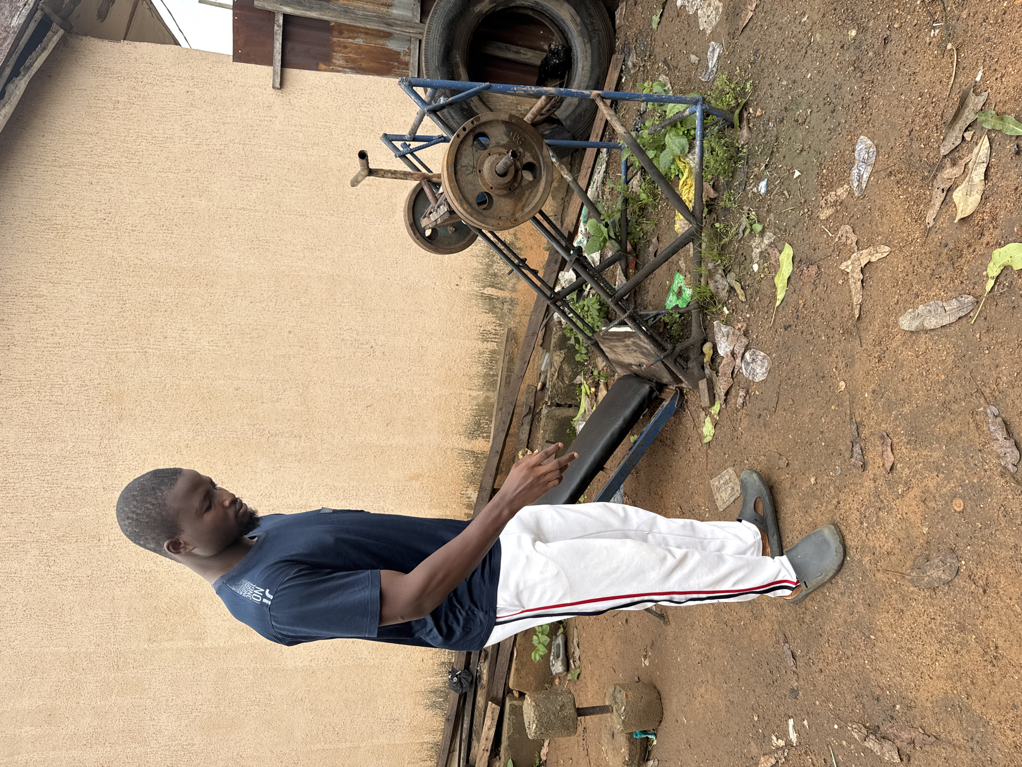 A young Black man in a dark blue t-shirt and white athletic pants with red and black stripes is positioned with his body rotated and facing left. He is standing next to a makeshift weightlifting bench with a blue metal frame. The bench is black and appears worn. In the background, a rusty metal weightlifting rack is visible with several weight plates. A large tire is propped up behind the rack. The ground is a mix of dirt, scattered leaves, and some debris, suggesting an outdoor, possibly cluttered, setting. A single concrete dumbbell is on the ground in front of the man. The overall scene appears to be an outdoor workout area. The lighting suggests it is daytime. The man's facial expression is neutral. There is some text visible on his t-shirt, but it is partially obscured and difficult to read. The location is Akwanga, Nigeria.