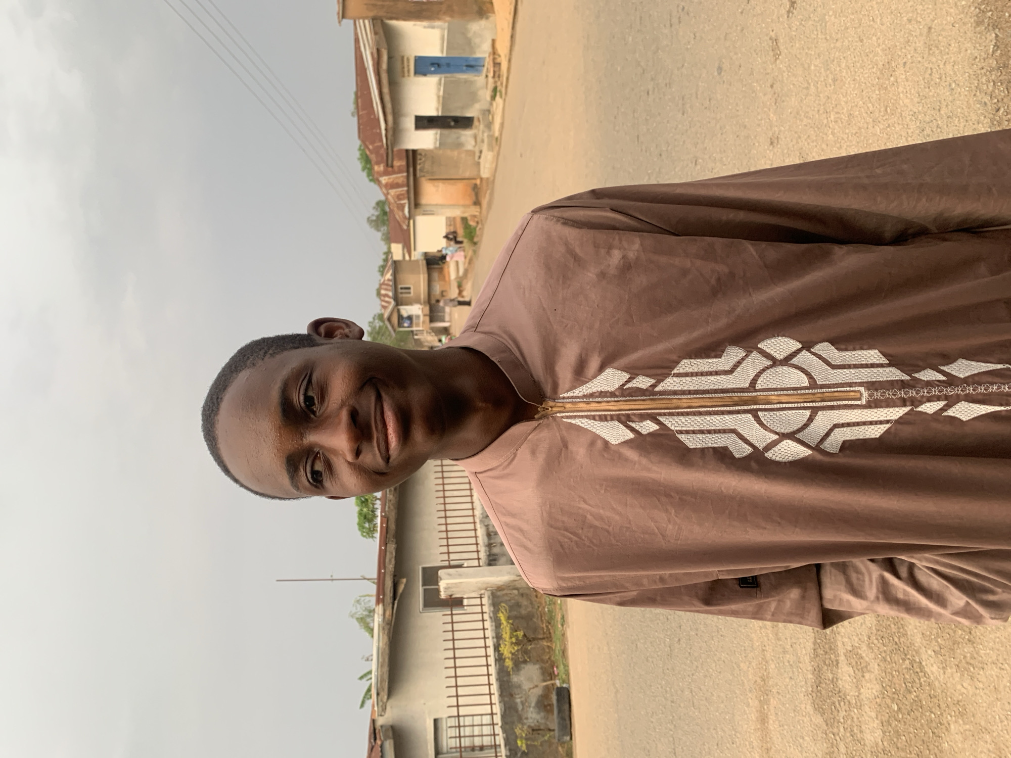A young man smiles at the camera while standing on a street in Jos, Nigeria. He wears a brown tunic with white geometric embroidery around the neckline. The street is paved and runs between buildings of modest construction, some with corrugated metal roofs. The sky is overcast. Power lines are visible overhead. Other people appear in the distance on the street.