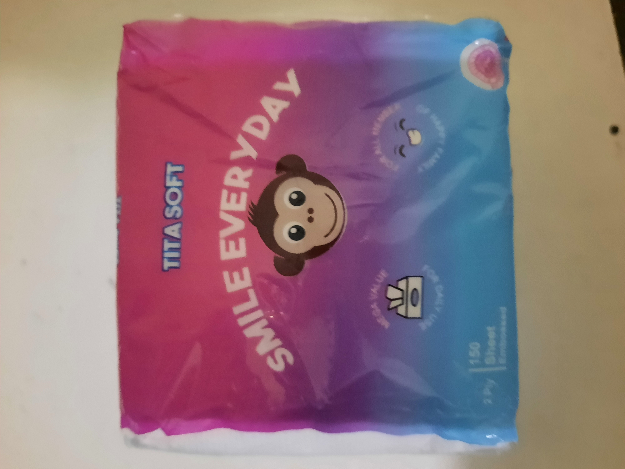 The image shows a package of TITA SOFT facial tissues, with a colorful gradient background of pink, purple, and blue. The brand name "TITA SOFT" is printed in blue at the top. The phrase "SMILE EVERYDAY" is printed in white across the center of the package, with a cartoon monkey face illustration. Below the monkey face, there are two circular graphics with text. The left graphic says "MEGA VALUE" with an illustration of a tissue box. The right graphic says "FOR ALL MEMBER" and "HAPPY FAMILY" with a smiling cartoon face. In the bottom left corner, it states "150 Sheet 2 Ply Embossed." The package is resting on a neutral-colored surface, possibly a table. There is a small pink circular sticker on the bottom right corner of the package. This image does not contain any information that would allow for its location in Lhokseumawe, Indonesia.