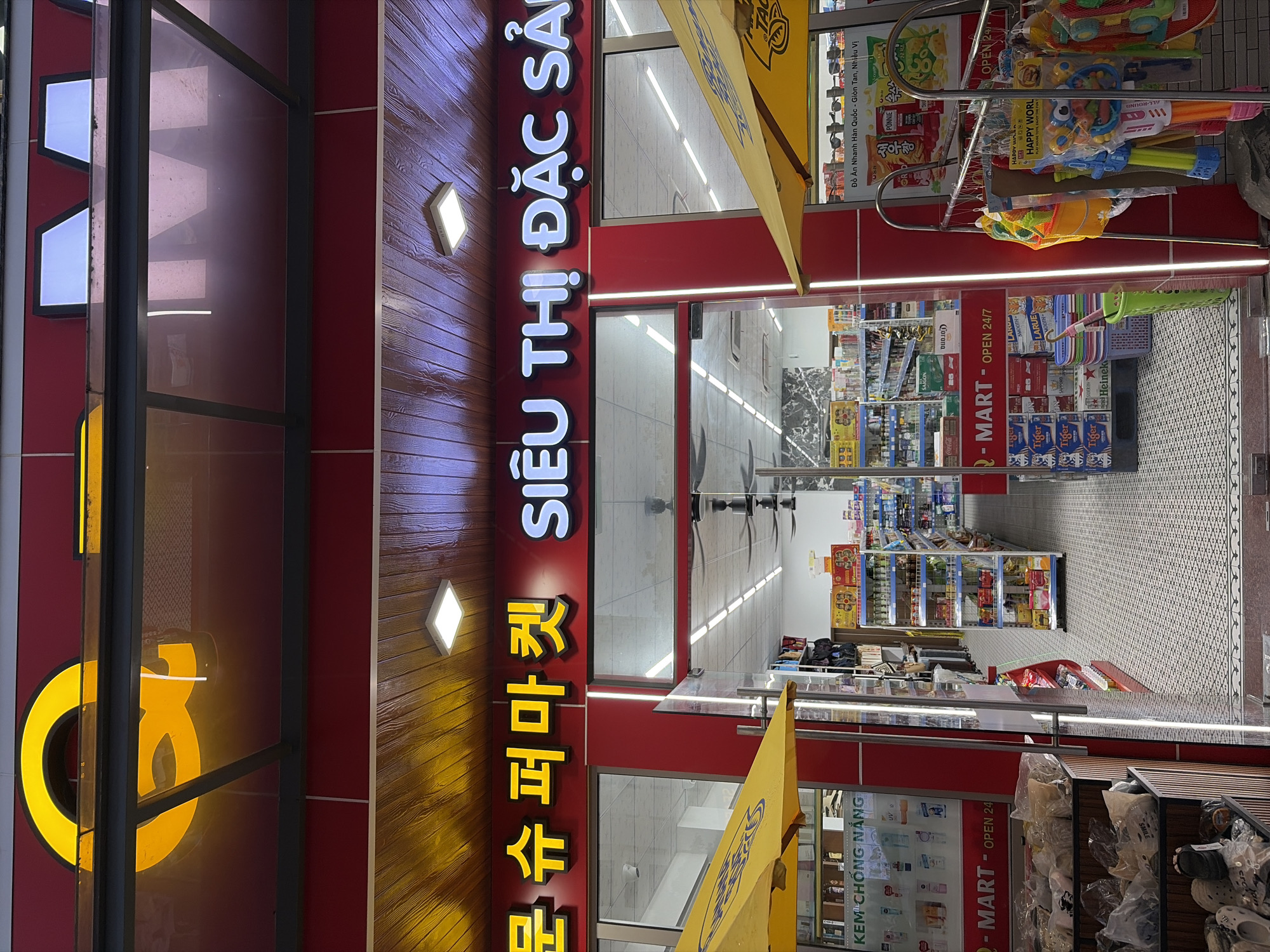 This is a street-level, medium-shot photograph of the exterior of a convenience store in Đà Nẵng, Vietnam. The store is identified by signage in both Korean and Vietnamese. The Korean text, in yellow, reads "문슈퍼마켓" (Munsyu Supermarket). The Vietnamese text, in white illuminated letters, reads "SIÊU THỊ ĐẶC SẢN" (Specialty Supermarket).
The store has a red facade with a wooden-paneled overhang above the entrance, illuminated by square ceiling lights. A large yellow awning is visible on the left, partially obscuring the store's interior and displaying Vietnamese text that reads "KEM CHỐNG NẮNG" (Sunscreen). Another yellow awning, partially visible on the right, also appears to be part of the store's branding.
The entrance to the store is open, revealing rows of shelves stocked with various products, including beverages, packaged goods, and cleaning supplies. Ceiling fans are visible inside the store. The floor of the store and the walkway outside are tiled with a patterned design.
In the foreground on the right, a rack displays children's toys, including water guns and buckets, with the text "HAPPY WORLD" visible on one of the packages. Bags of merchandise are stacked in wooden bins to the left of the entrance. The overall impression is a clean and well-stocked convenience store ready for business.