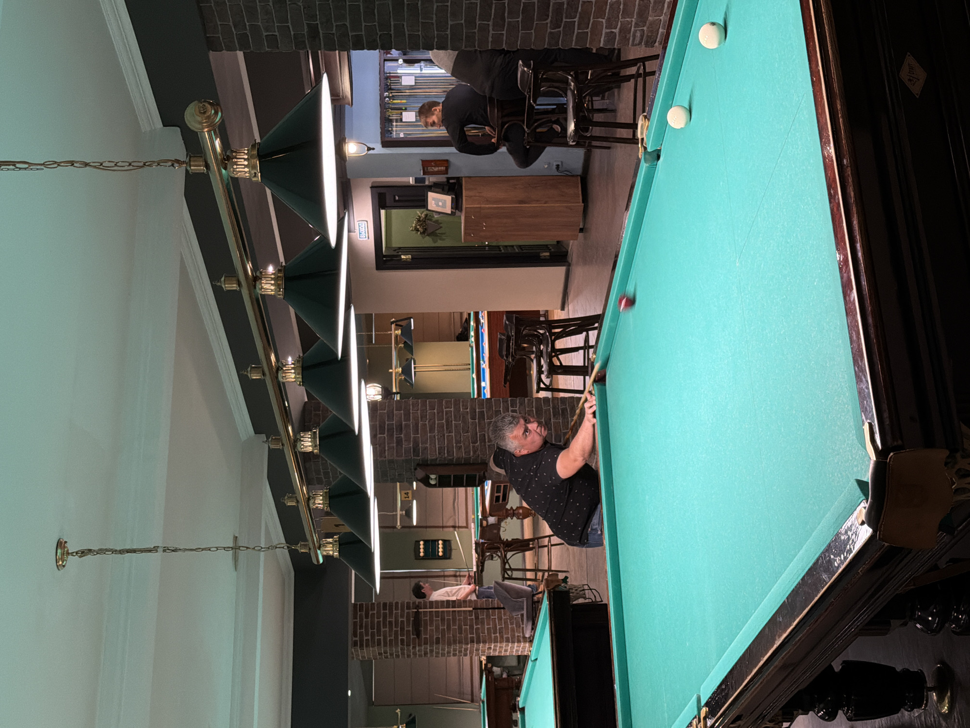 This image depicts an indoor scene within a billiard club in Izhevsk, Russia. In the foreground, a middle-aged man with short, graying hair, wearing a dark polo shirt with light patterns and blue jeans, is actively engaged in a billiard game. He is bent over a large, green-felt billiard table with ornate dark wood legs, holding a cue stick, and appears to be in the process of making a shot, focused on a red ball near a pocket, with two white balls also visible on the table.
The club features multiple billiard tables extending into the background. To the mid-left, another man in a white t-shirt stands at a separate billiard table, holding a cue. Towards the right, two men are seated on high stools; one, dressed in a dark long-sleeved shirt, faces left, observing the main player. The club's interior is characterized by dark brick-clad pillars and walls, a dark floor, and a white ceiling with decorative crown molding. A series of brass-colored linear chandelier fixtures with multiple dark conical shades hang from the ceiling, providing primary illumination. In the background, a dark green door with a white "ВЫХОД" (EXIT) sign is visible next to a wooden cabinet, and wall-mounted lamps provide additional ambient light. Cue racks and other billiard tables are dimly visible further in the background.