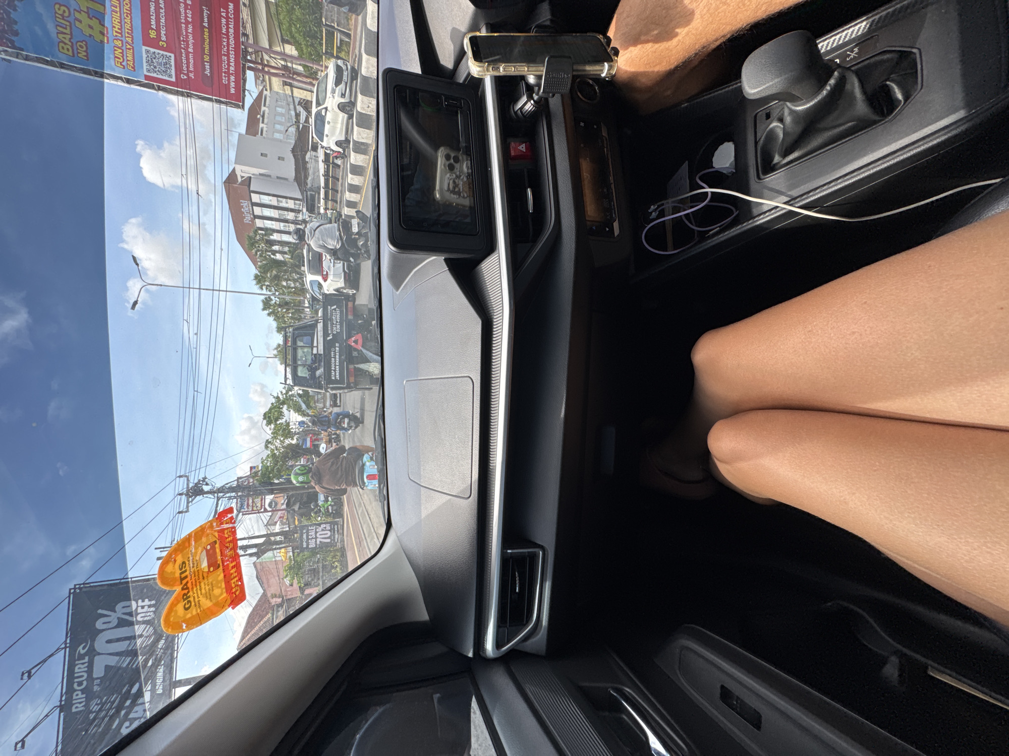 The image shows an interior car view looking out onto a street in Kuta, Indonesia. The dashboard and center console of a vehicle are visible, with a phone mounted on the dash. The view through the windshield reveals street activity, including cars, motorcycles, and pedestrians. A large "RIPCURL 70% OFF" advertisement and another ad for "BALI'S NO #1 FUN & THRILLING FAMILY ATTRACTION" are visible. Fairfield is noted in the background. A pair of legs, apparently belonging to a passenger, are in the foreground.