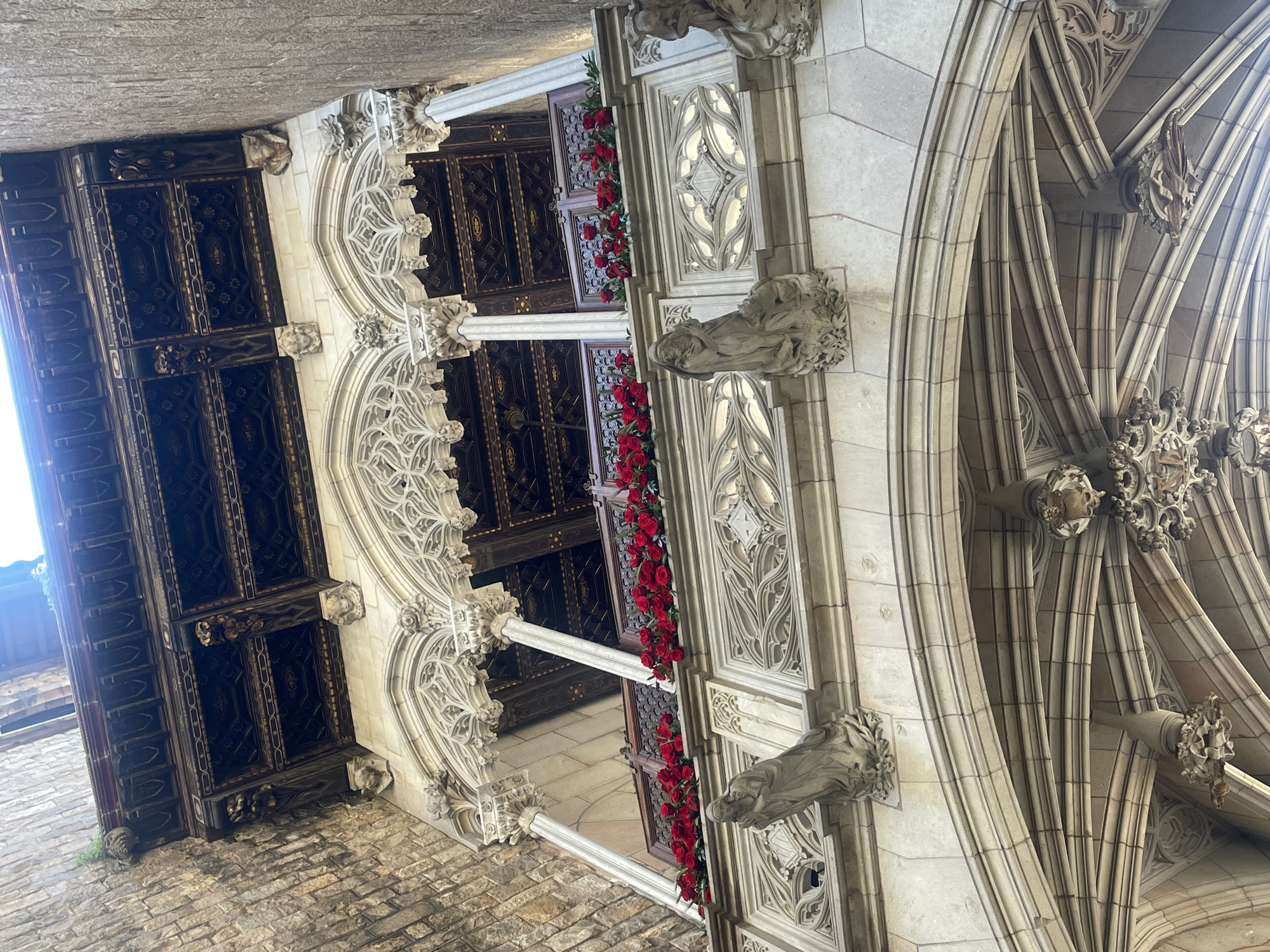 This media file captures a detailed view of ornate Gothic architecture, likely within a cloister, courtyard, or grand hall in Barcelona, Spain. The image is taken from a low angle, looking upwards at a convergence of architectural elements.
On the right, a complex system of light-colored stone ribbed vaulting creates an intricate ceiling or upper wall, adorned with decorative bosses and carved elements. Below this, sculpted gargoyles or grotesques project from the stone work, adding to the historic aesthetic.
To the left, a series of dark, richly paneled wooden structures, possibly windows or doors, are set into an elaborately carved stone facade. These wooden elements feature intricate geometric and possibly floral patterns. Along the base of these openings, vibrant red roses are arranged in long, neat rows, providing a striking splash of color against the otherwise monochromatic stone and dark wood. Intricate stone tracery is visible in the window openings above and around these wooden sections. Further to the left and lower, a rough, stone brick wall forms the foreground, suggesting an outdoor or semi-enclosed space.
The scene is illuminated by bright natural light, suggesting it is daytime. There are no people visible, and no specific activity is taking place, allowing the focus to remain on the architectural grandeur and craftsmanship. The combination of detailed stone carvings, wooden elements, and the contrasting red roses creates a sense of historical elegance and dramatic beauty. No visible text is present in the image.