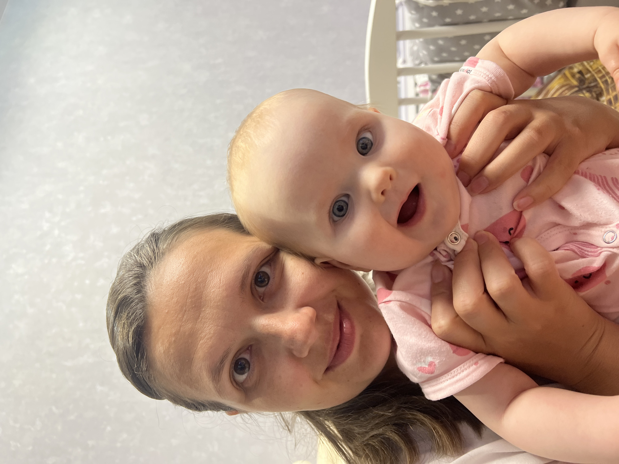 The scene is an indoor setting, consistent with a residence, located in the city of Chaikovsky, Russia. The image features two individuals: an adult female and an infant, captured in a close-up, chest-up shot.
The adult female, positioned on the left, has dark brown hair pulled back, with visible strands across her forehead. Her eyes are dark brown, and she looks directly at the camera with a soft, closed-mouth smile. She appears to be wearing a light-colored garment, possibly white or light pink, visible on her left shoulder and arm. Her hands are wrapped around the infant.
The infant, positioned on the right and held by the adult, has light blonde hair and wide, light blue eyes, also looking directly at the camera. The infant's mouth is slightly open. The infant wears a pink long-sleeved onesie adorned with various lighter and darker pink patterns, including small hearts. Snap closures are visible on the front of the onesie. The adult's hands are visible holding the infant's torso, with fingers wrapped around the infant's upper body and arm.
The background consists of a light-colored (off-white or very light gray) wall with a subtle, textured pattern. To the right of the infant, a section of a white crib railing is visible, behind which a gray and white patterned fabric or bedding (possibly with star patterns) can be discerned. The lighting is even and appears to be artificial indoor illumination.
