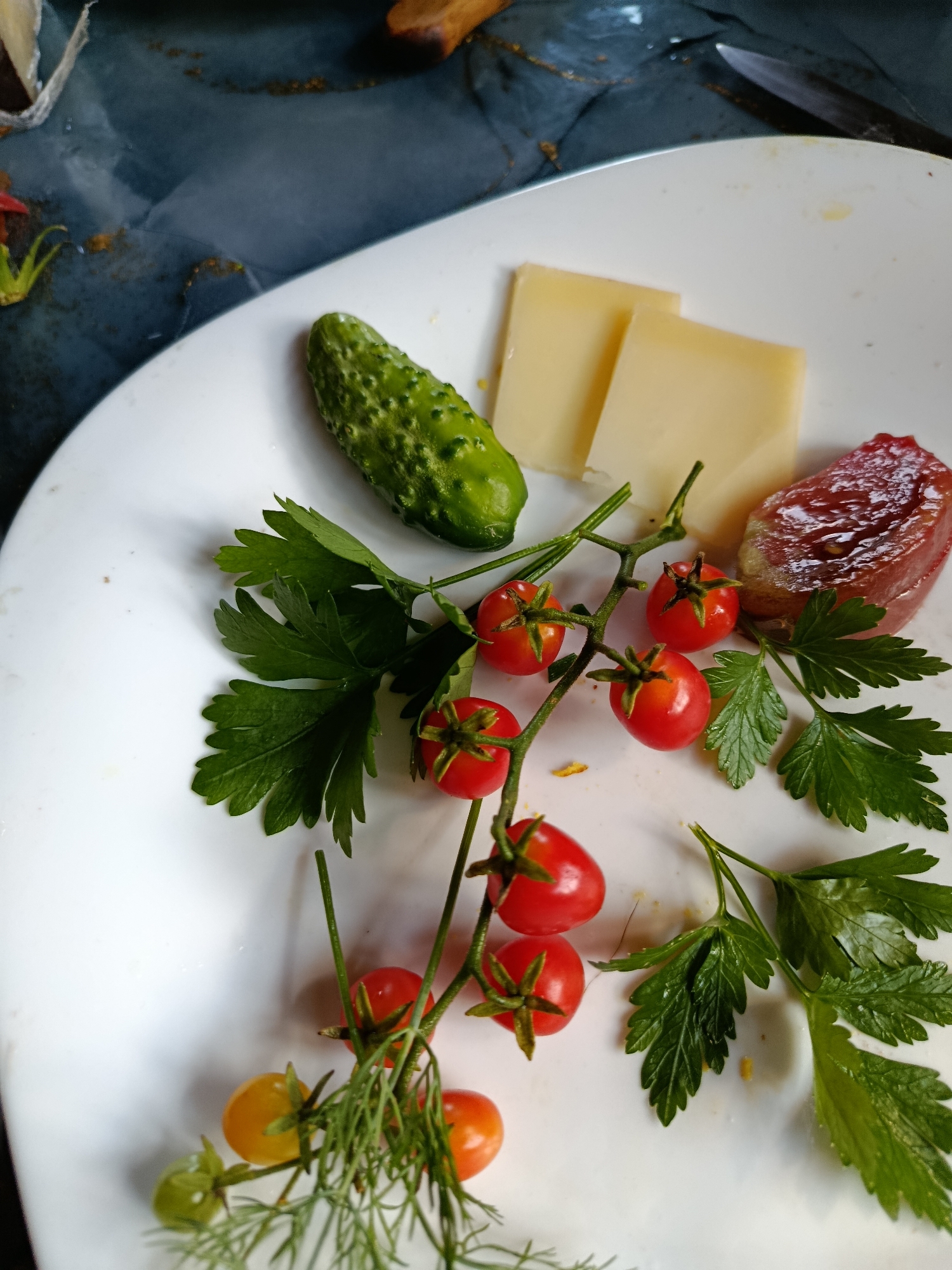 A close-up shot showcases a white plate with a small collection of appetizer items. Positioned on the plate are a single, small, bumpy cucumber, two thin slices of pale yellow cheese, and a slice of cured meat that is dark red and slightly translucent. A sprig of cherry tomatoes, with most of the tomatoes a vibrant red and a few transitioning to yellow and green, is fanned out across the plate. Fresh green parsley sprigs are scattered amongst the tomatoes. The background is a dark, marbled surface, possibly a countertop or table, with some hints of gold flecks. A glint of a metal object, possibly a knife, is visible in the upper right corner. There are no people or actions depicted. This scene is set on a plate, suggesting a prepared meal or snack. The specific location in the city of Верея, Russia, is not discernible from the visual information provided in the image.