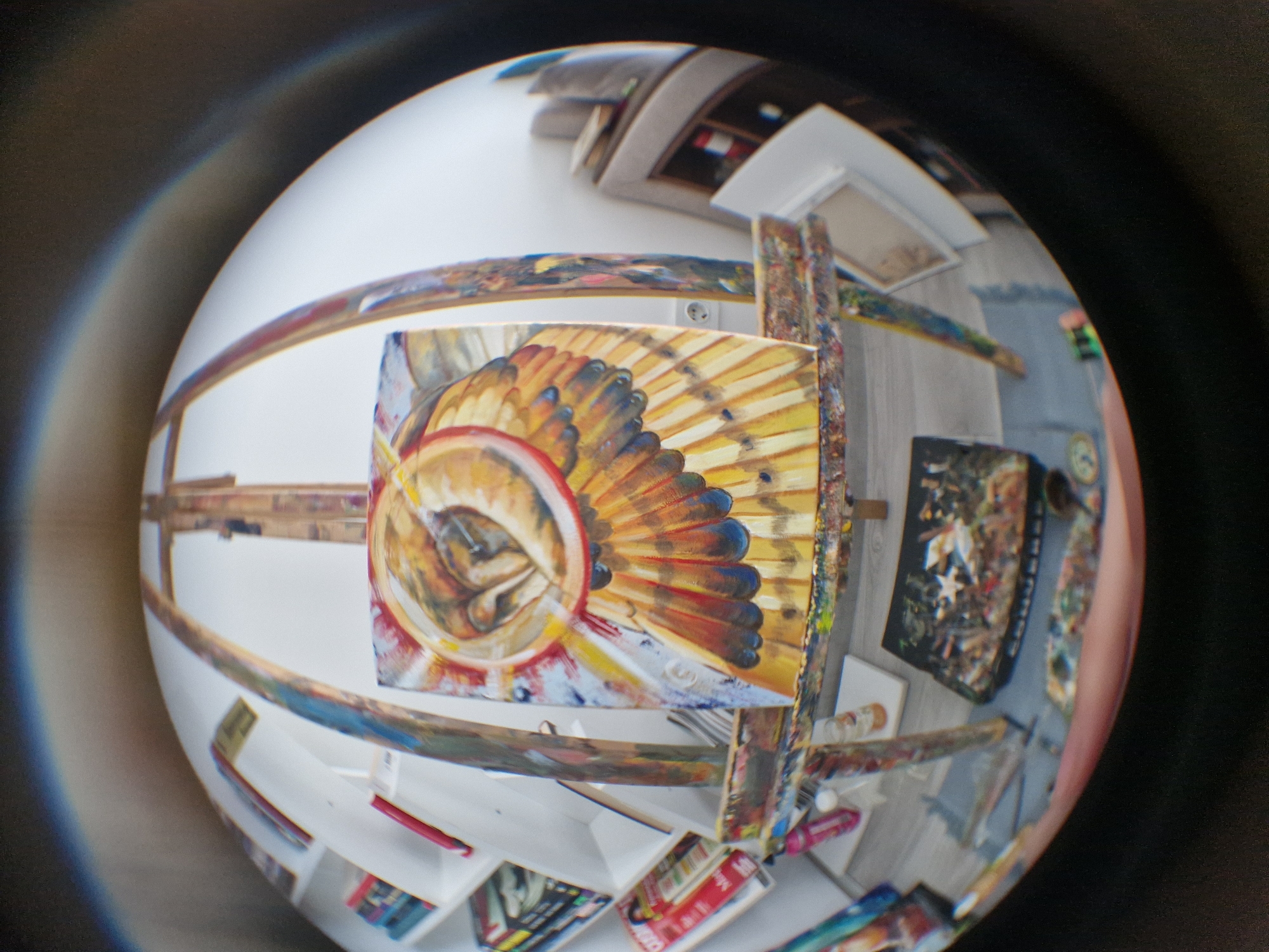 A fisheye lens photograph captures a vibrant artist's studio, dominated by a colorful painting on an easel. The painting features an abstract or stylized design resembling feathers or a headdress, with a prominent circular motif in the center, rendered in shades of yellow, orange, red, and blue.
The wooden easel supporting the canvas is visibly splattered with various paint colors, indicating active use. Surrounding the easel, the distorted background reveals elements typical of an art workspace: shelves filled with art supplies, books, and other canvases. Paint tubes, brushes, and various art materials are scattered across surfaces and the floor. A white power outlet is visible on a wall.
The scene suggests an ongoing creative process within an indoor setting. The lighting appears to be bright and evenly distributed. Due to the fisheye distortion, the exact dimensions and layout of the studio are warped, creating a spherical view of the room.
Visible text on some items in the background includes "Made in", possibly "CHINA", "BEST", and "COLOR", suggesting labels on art supplies or packaging. No people are visible in the image, and the time of day or weather cannot be determined. The overall impression is one of an active and colorful artist's environment.