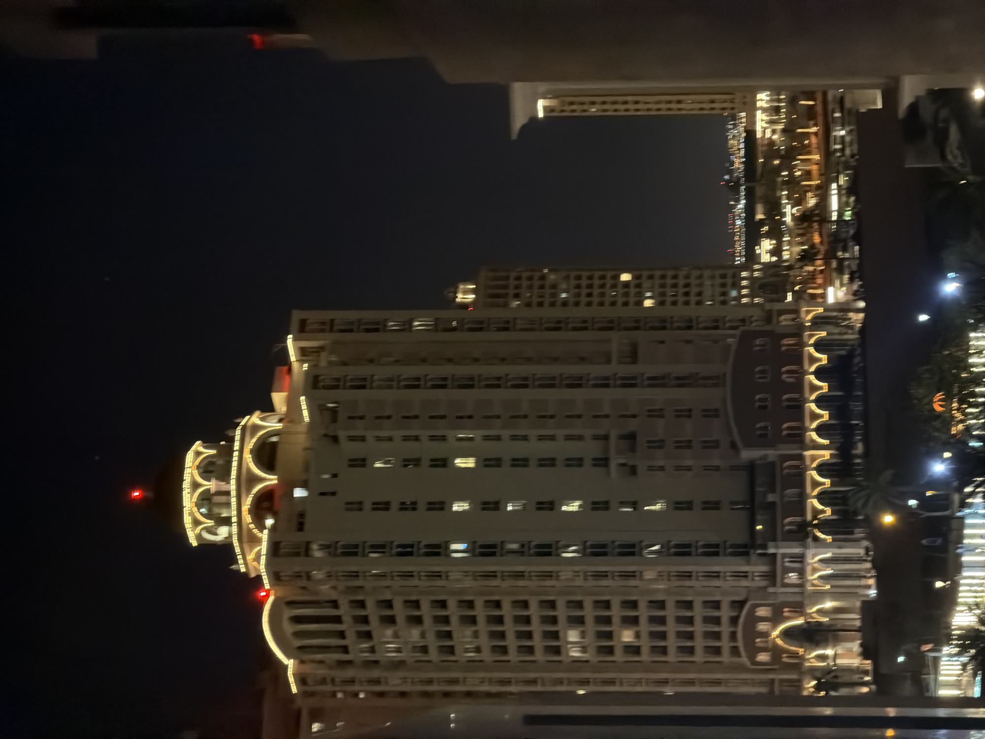 An exterior, eye-level night photograph captures a prominent, multi-story building featuring classical architectural elements within an urban setting. The building is light-colored, possibly beige or off-white, and extensively outlined with warm yellow-orange decorative lighting that highlights numerous arches on its lower and upper floors. A large central dome tops the structure, also illuminated by decorative lights and capped with a red aviation warning light. A second red light is visible on a lower roof section to the left. Numerous windows are visible, some illuminated from within with yellow-white light, and one distinct blue light on a central upper floor. Palm trees, dimly lit by ground-level illumination, are discernible at the building's base. Further into the mid-ground, additional city lights and structures are present. The background consists of a dark, clear night sky with faint distant lights. Blurred sections of adjacent buildings or structures frame the image on the left and right, with a thick, dark column visible on the far right. No individuals or discernible actions are present. The scene depicts a modern urban environment at night. The architectural style and prominent dome suggest the building is located in The Pearl-Qatar, Doha, Qatar.
