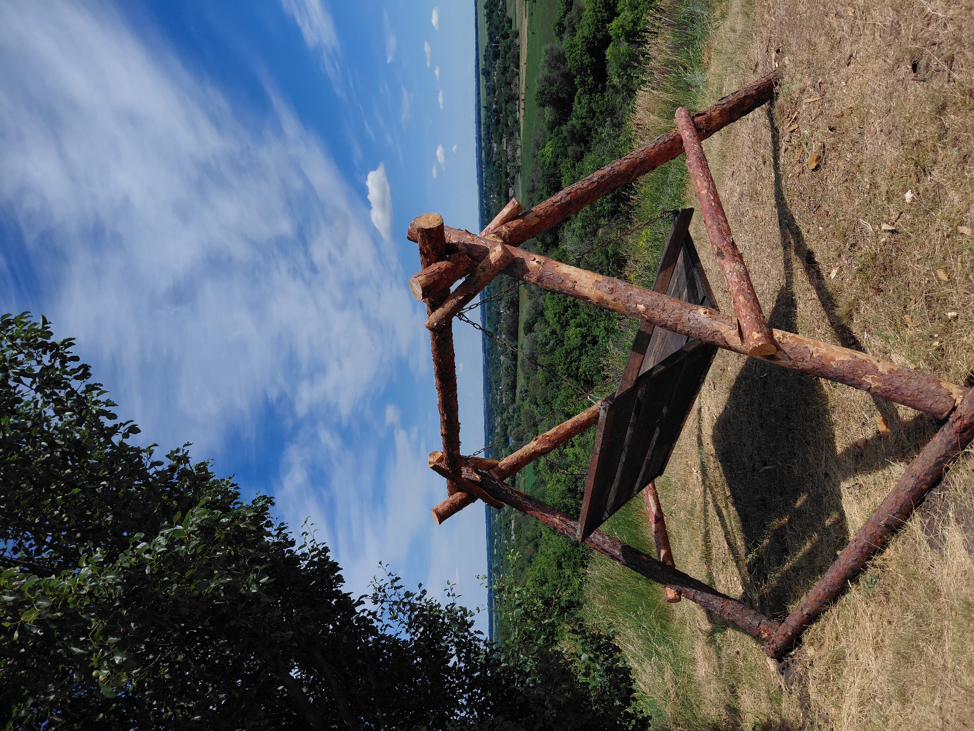 A rustic wooden swing is set against a backdrop of rolling green hills and a bright blue sky with wispy white clouds. The swing, constructed from rough-hewn logs, is positioned on a grassy knoll, casting a shadow on the dry ground. In the foreground, the dense green foliage of a tree partially obscures the sky and the upper portion of the swing. The middle ground features a panoramic view of a lush, verdant landscape dotted with trees and some scattered buildings, indicative of a rural or semi-rural setting. The overall scene evokes a sense of tranquility and natural beauty.