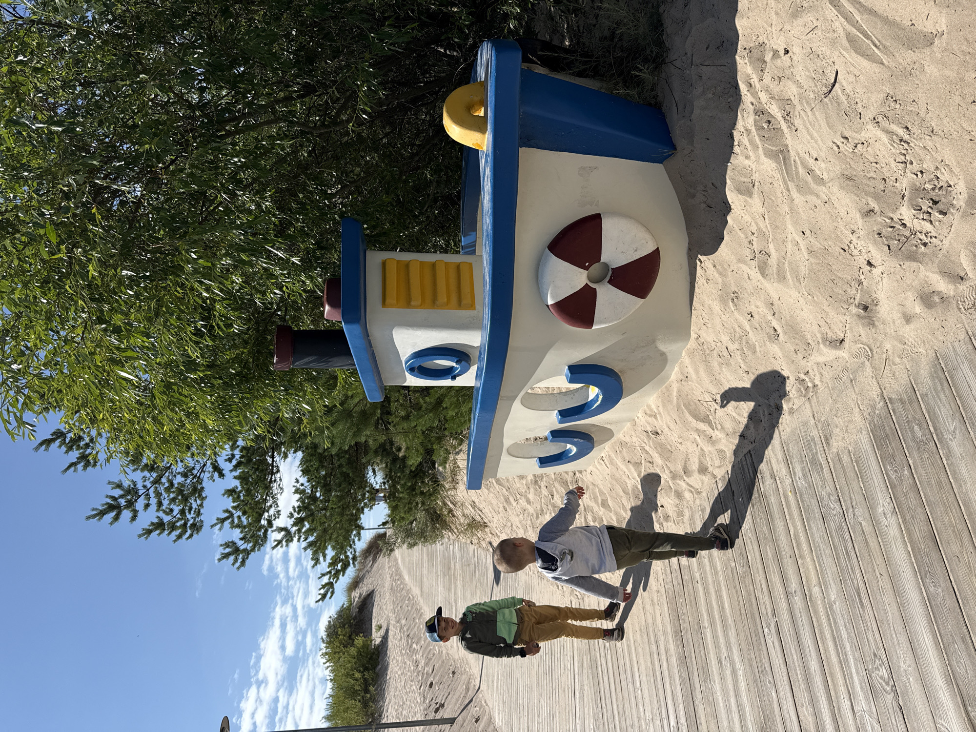 Two young boys, one wearing a green and grey hooded sweatshirt and tan pants, and the other in a grey sweatshirt and green pants, are walking on a wooden boardwalk. They are in Ventspils, Latvia, at the children's town nautical-themed park. To their right, a large, white, blue, and yellow toy boat is situated in the sand. The boat has a red and white life preserver attached to its side and a yellow ring on its deck. Lush green trees and shrubs form a backdrop, with a clear blue sky above. The sun casts shadows of the boys and the boat onto the sand.