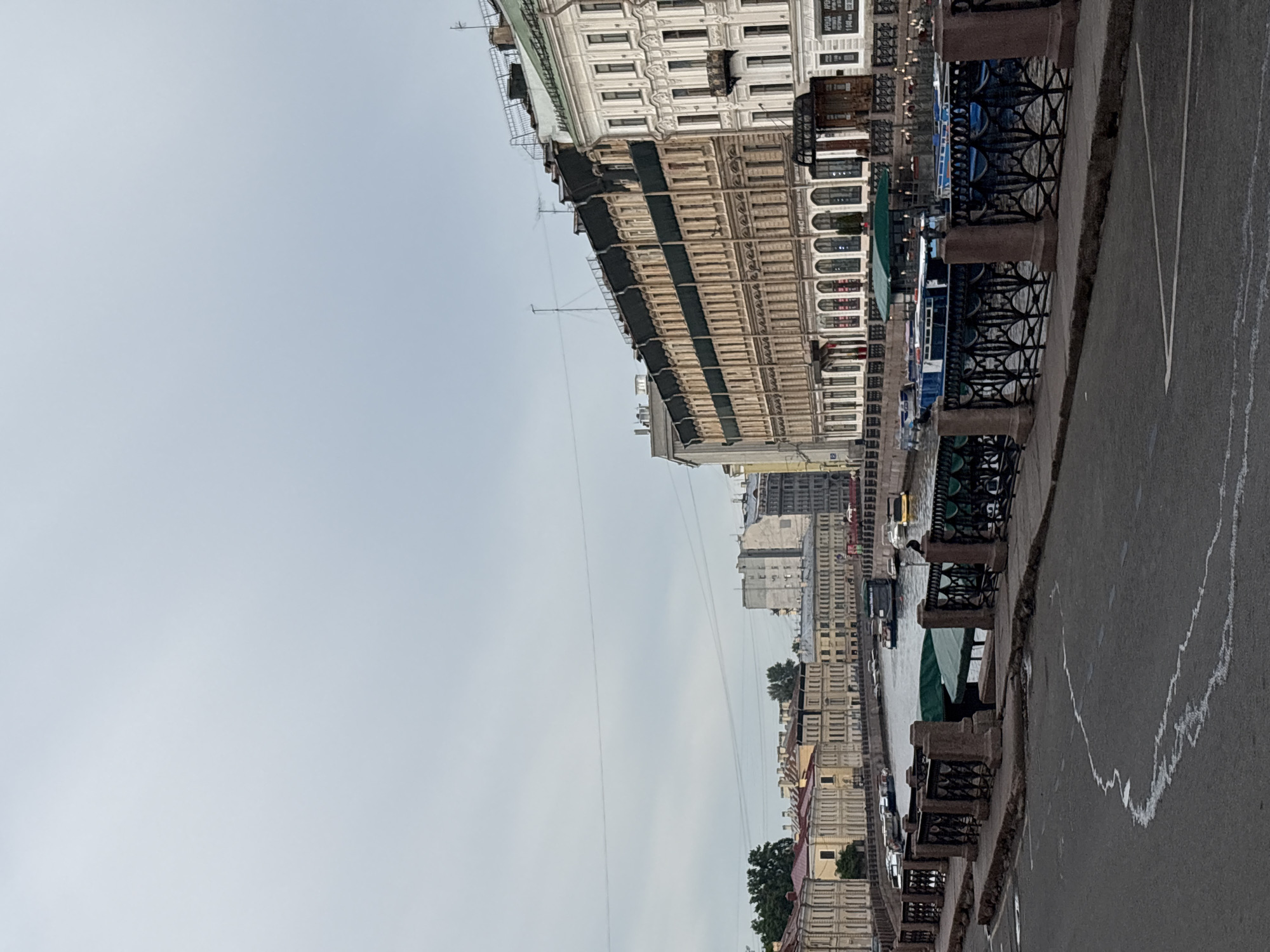 The image shows a canal in Saint Petersburg, Russia, with buildings lining its banks. The canal is lined with boats, and there is a walkway and a road in the foreground. The sky is overcast, and the buildings appear to be old and historic, with ornate architecture. The scene is one of a typical European city canal.