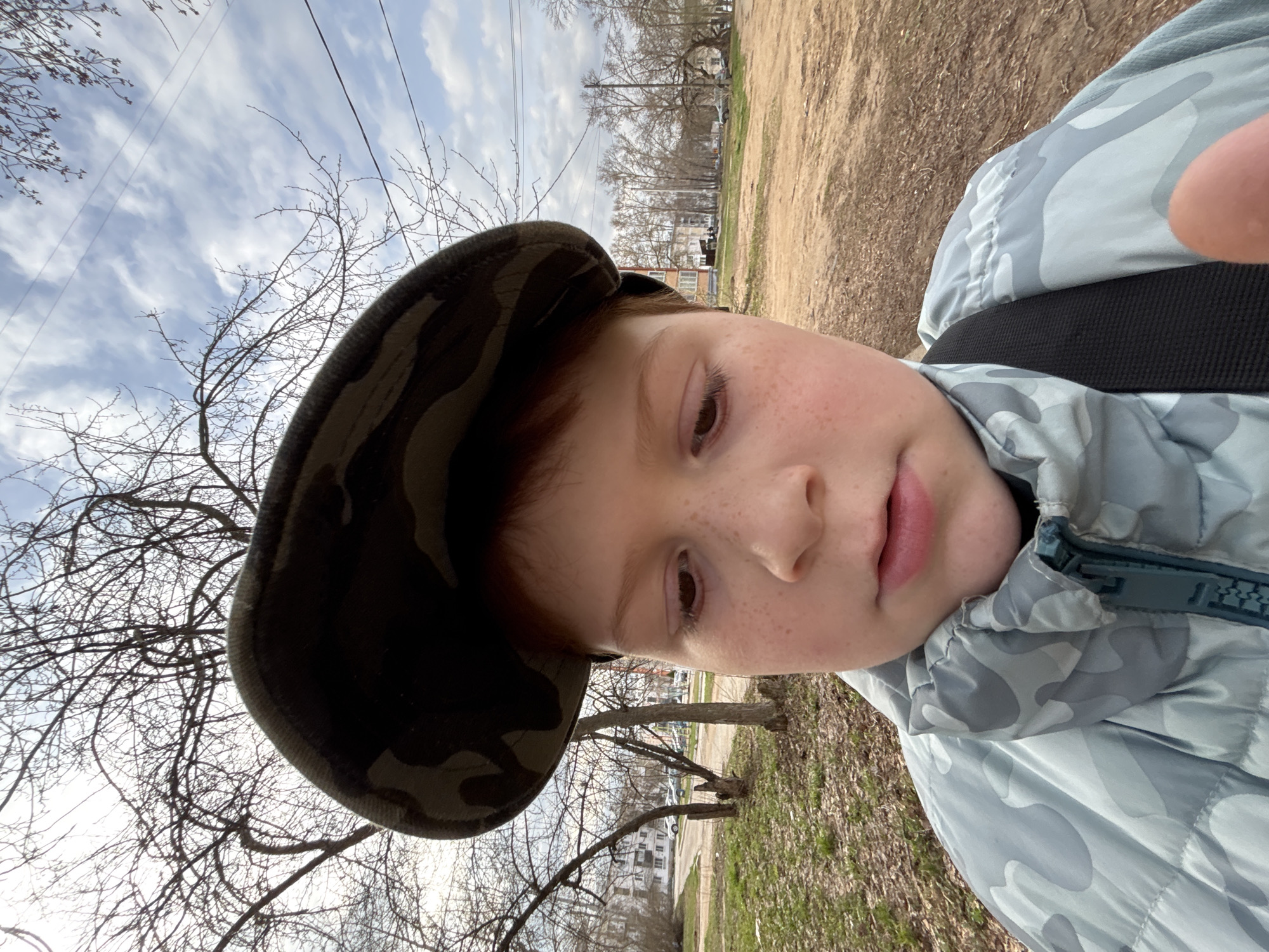 A young child, likely male with fair skin and freckles, wears a camouflage patterned hat and jacket. The child appears to be taking a selfie, as part of a finger is visible in the upper right. The background includes bare trees, a dirt path, buildings, and a cloudy sky. Power lines are visible overhead. Based on the style of architecture and infrastructure, the location is likely Chaikovsky, Russia.