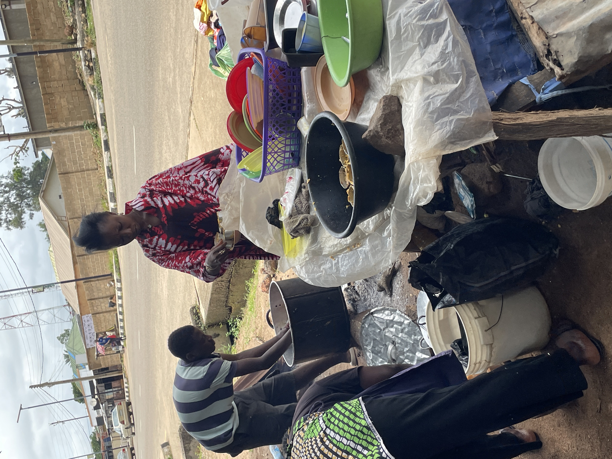 This image captures an outdoor scene on a street in Jos, Nigeria, likely a market or roadside stall. In the foreground, a woman in a vibrantly patterned red and white garment stands over a collection of plastic bowls and plates. She appears to be engaged in an activity related to food preparation or serving, holding a small bowl in her left hand.
To the left, a man wearing a striped t-shirt and dark shorts is hunched over a large, black pot, his hands submerged in it, suggesting he is washing or preparing something. Another person, partially visible in the lower left corner, wears a green and yellow patterned vest over dark clothing.
The ground around them is a mix of dirt and some scattered debris, including plastic bags, rocks, and metallic objects. A white bucket and a large black plastic bag are also visible. The background shows a paved road with cars and a sidewalk. Buildings with brick walls and some vegetation are present, along with a prominent radio tower in the distance, a common landmark in many Nigerian cities. The sky is overcast, indicating a cloudy day. The overall impression is of a bustling, everyday scene of commerce and activity on a Nigerian street.