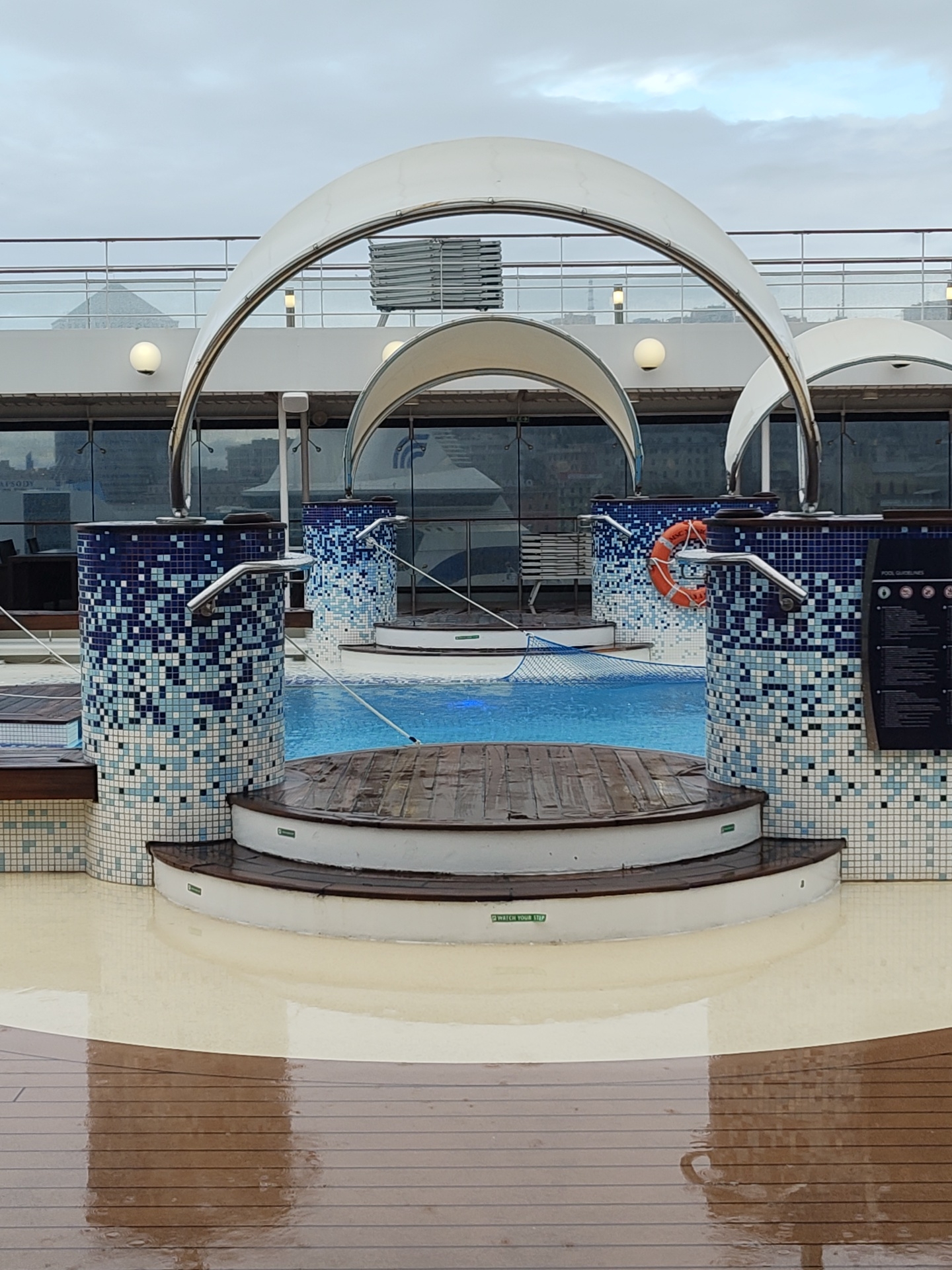 The image shows a pool area on a cruise ship. The pool is surrounded by mosaic-tiled pillars and a wooden deck. Above the pool are white arches. A life preserver hangs on one of the pillars. In the background, windows reveal a view of Genoa, Italy.