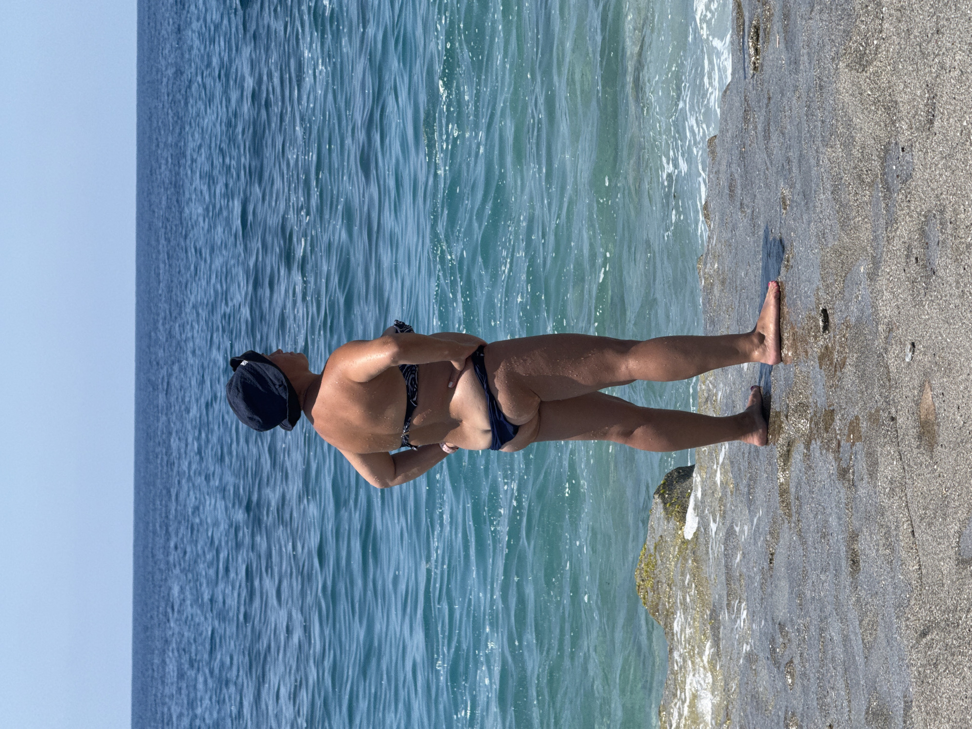 An adult female, observed from behind, stands on a wet, dark grey, textured coastal platform in Alanya, Turkey. She wears a dark blue or black bucket hat, a patterned dark bikini top, and dark bikini bottoms, with her hands resting on her hips. Her skin glistens with water droplets, and her left toenail is painted red. She faces the expansive blue sea, which exhibits a gradient from clear turquoise near the shore to a darker blue towards the distant horizon. Gentle waves break against the platform, and a rock partially covered with green algae is visible to the subject's left. The sky above is clear and light blue, indicating fair weather.