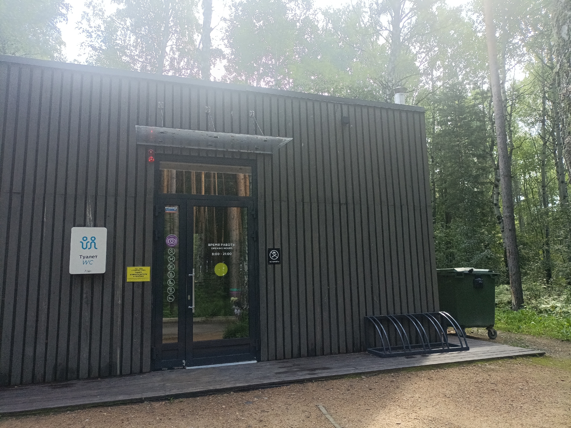 The image depicts an exterior view of a modern public restroom facility situated in a wooded area. The building has a dark gray, vertical wooden plank exterior. A glass-paned door with a metal handle provides entrance. Above the door, a metal awning is visible.
To the left of the entrance, a white sign displays the universal symbol for a toilet and the word "Туалет" (Toilet) in Russian, along with "WC". Below this sign, a smaller yellow sign reads "ВРЕМЯ РАБОТЫ OPENING HOURS 8:00 - 21:00". A vertical row of circular symbols is affixed to the doorframe, to the right of the entrance. One symbol appears to be a camera, and others depict various icons. A black circular sign with a white cigarette and a diagonal line through it, indicating "no smoking", is mounted on the building to the right of the door.
In front of the building, a wooden platform extends outwards. A black metal bicycle rack, capable of holding several bicycles, is positioned on this platform. To the right of the platform and building, a large green dumpster with wheels is visible.
The background consists of a dense forest with tall trees, suggesting the location is in a natural or park-like setting. The lighting suggests it is daytime, with some atmospheric haze or glare present. The ground in front of the building is a mix of gravel and dirt.