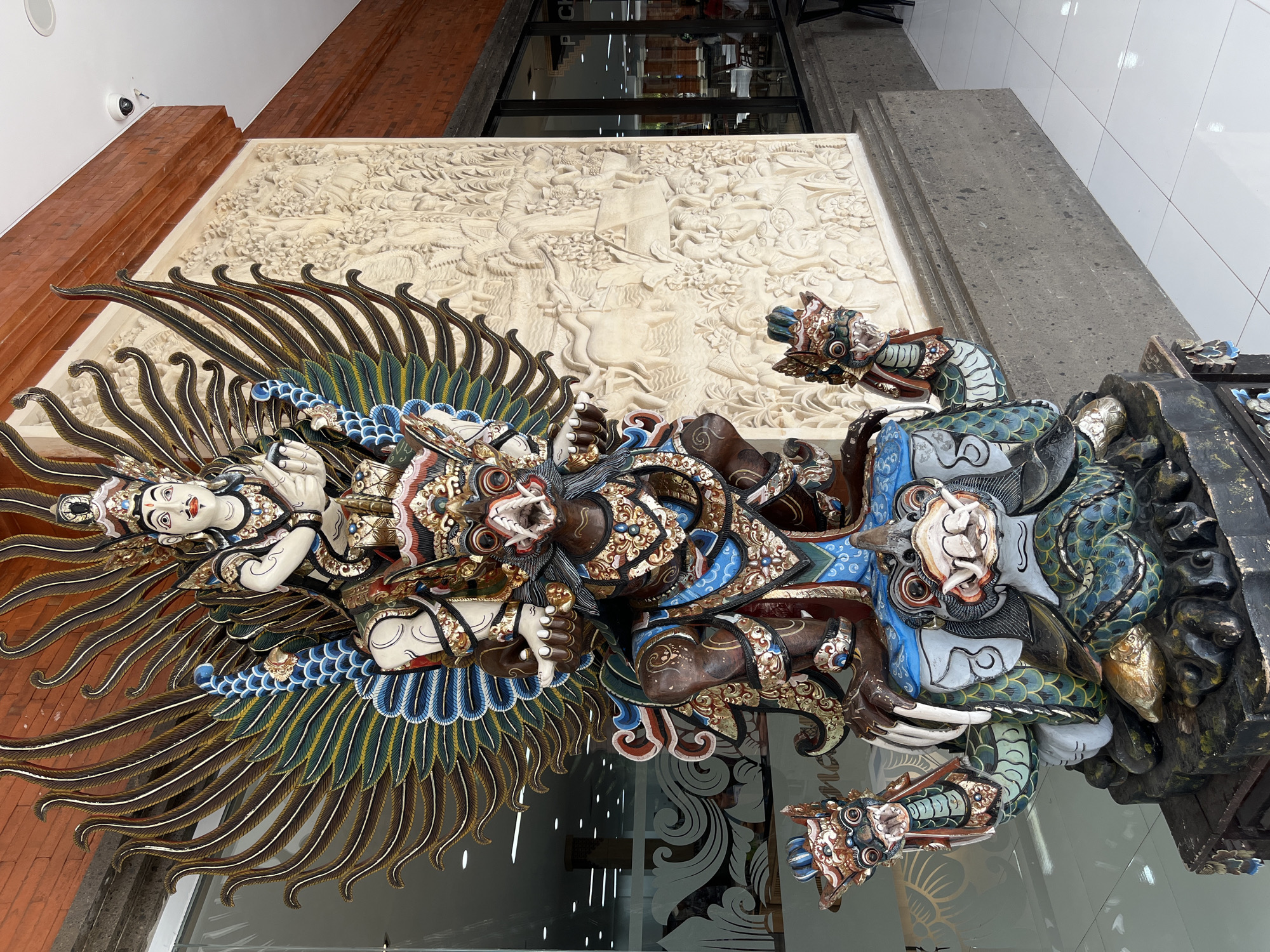 This is a medium shot, eye-level, outdoor photograph of a large, elaborately carved and painted wooden statue in Kuta, Indonesia. The statue depicts a mythological figure with multiple heads and wings, possibly Garuda. The statue is predominantly dark brown and blue, with gold accents, and is adorned with intricate patterns and detailing. It is positioned in front of a textured wall with bas-relief carvings of figures and animals, likely depicting a scene from Balinese mythology or daily life. To the left of the statue is a glass-fronted building with reflections of lights and interior structures visible, suggesting a commercial establishment such as a shop or restaurant. To the right, a portion of the building's exterior, clad in brick, is visible, along with large windows offering a glimpse into a seating area. The ground is tiled with white squares. There are no people clearly visible in the foreground or immediate surroundings of the statue. The lighting suggests it is daytime.