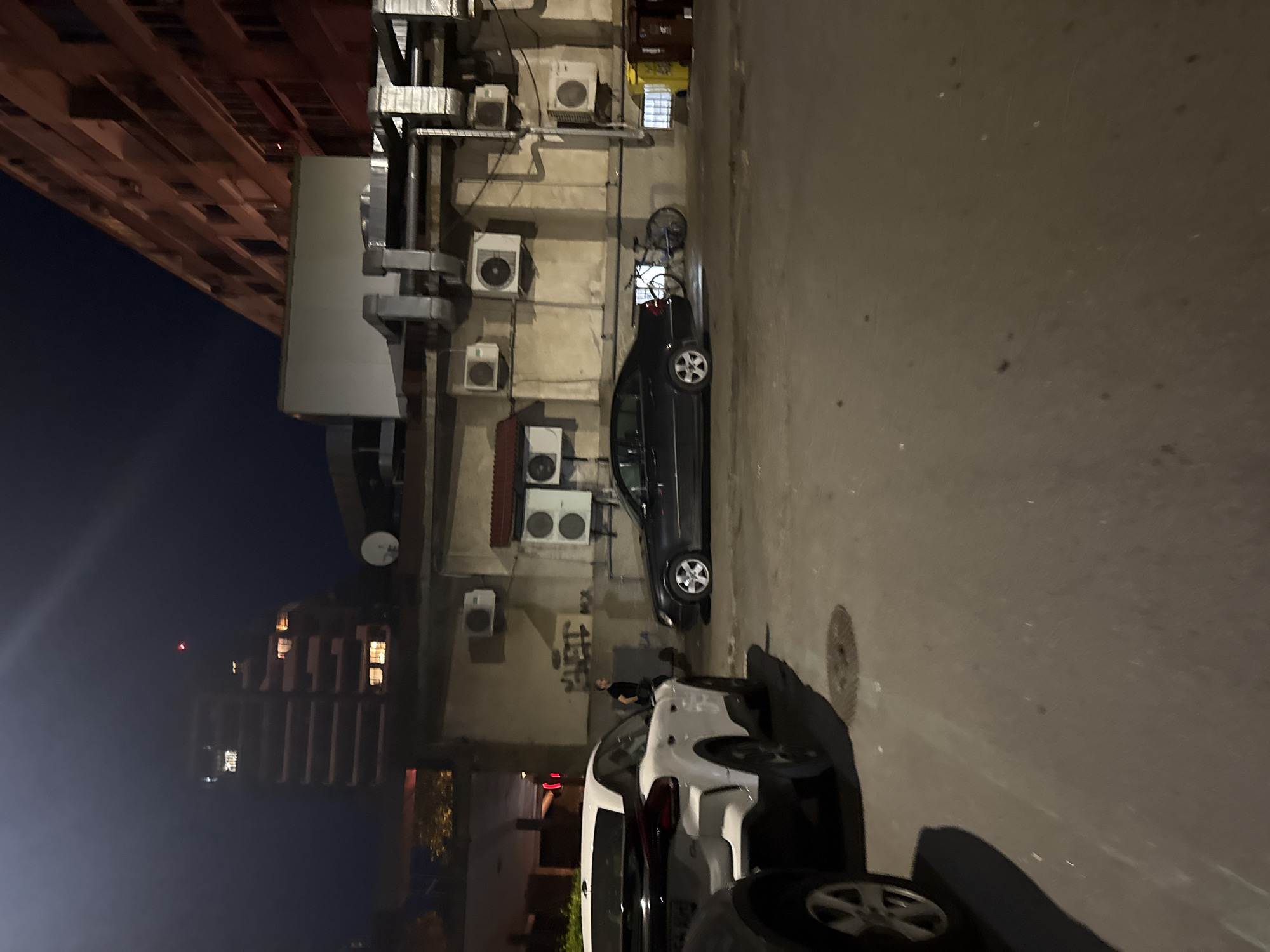 An outdoor nighttime scene in an urban environment, possibly Burgas, Bulgaria, shows several parked cars on a paved surface. In the foreground, the rear of a white SUV is visible on the left. To its right, a dark-colored sedan is parked. A bicycle is positioned behind the sedan. A person in a dark shirt stands near the vehicles. The background features a multi-story building with numerous windows and visible air conditioning units attached to the facade of a lower structure. There are also trash receptacles visible. The sky is dark.