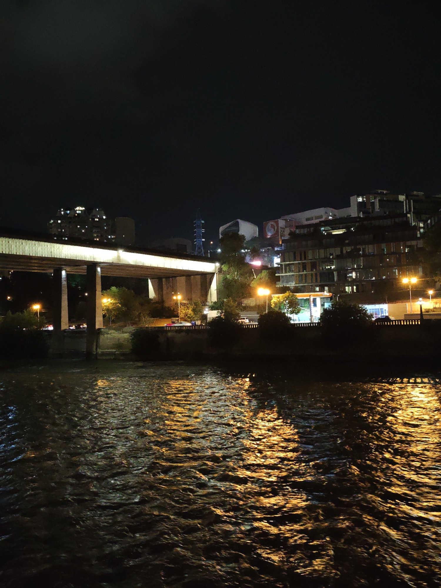 This is a nighttime photograph of a wide river flowing through the city of Tbilisi, Georgia. The water's surface is choppy and reflects the warm glow of artificial lights from the city. In the background, a large overpass or bridge, illuminated from below, stretches across the scene. Beyond the bridge, the cityscape rises, featuring several buildings with illuminated windows, suggesting residential or commercial spaces. One prominent building, possibly an apartment complex, has a distinctive pattern of lights. Further back, a tall, blue-lit structure, possibly a power transmission tower, is visible against the dark sky. The overall scene conveys a sense of urban activity at night, with the river acting as a central element reflecting the city's illumination.