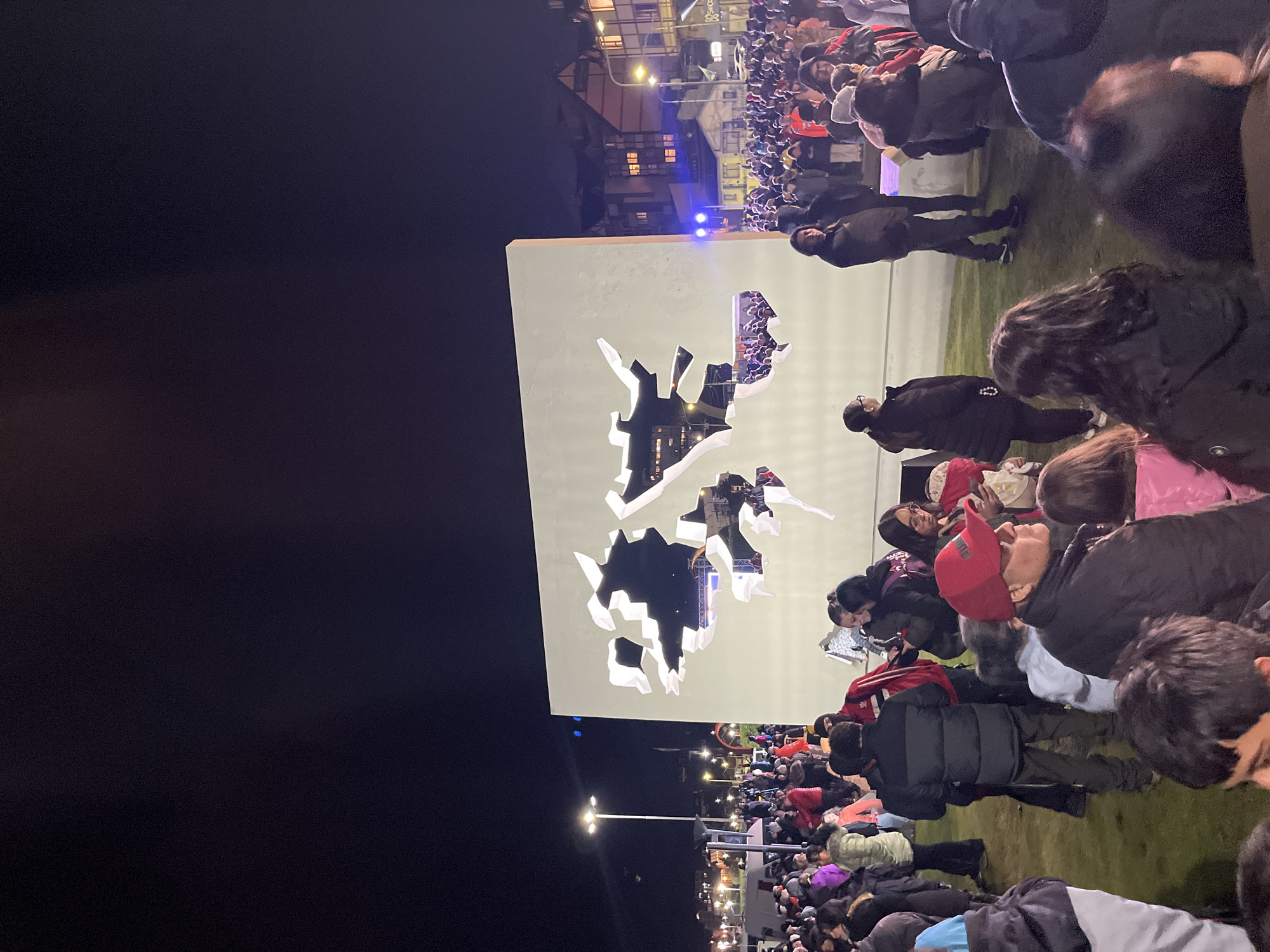 At night in Ushuaia, Argentina, a crowd gathers on a grassy area in front of a large white structure featuring cutouts that resemble the shape of the Falkland Islands. People are mostly in winter clothing, and some are wearing hats. Streetlights illuminate the scene, and buildings can be seen in the background.