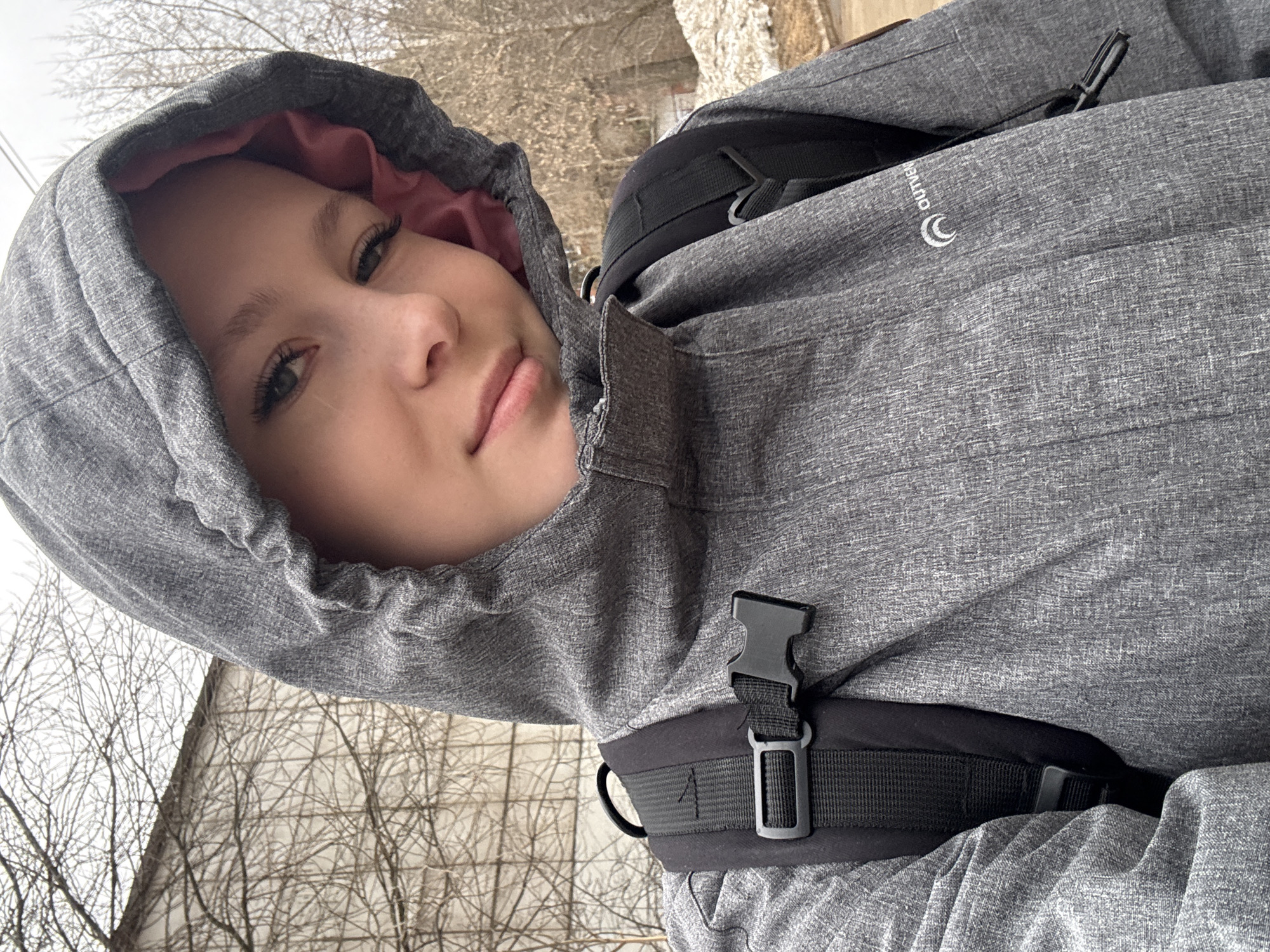 A young person, possibly a woman, with fair skin and visible eyelashes, is shown wearing a gray hooded jacket with a pink lining. The hood is up, framing their face. The person is also wearing a black backpack, as indicated by the straps and buckle visible on their chest. The jacket has the brand "OUTVER" displayed on the upper right chest.
The background reveals leafless tree branches and a building partially covered with snow, suggesting a winter or early spring season. The setting appears to be outdoors, possibly a city or suburban environment.
Based on the metadata, the location is Chaikovsky, Russia.