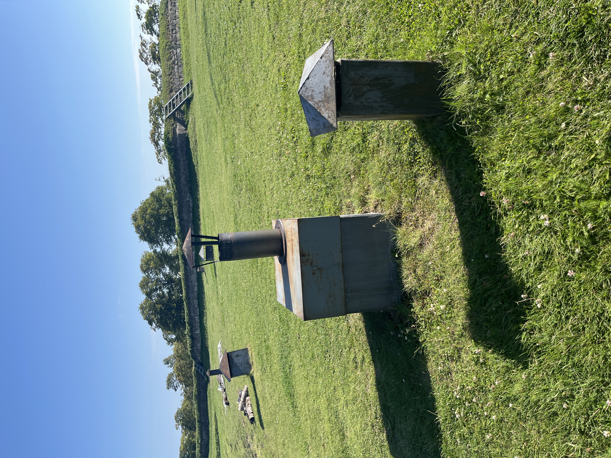 The image depicts an outdoor scene on a sunny day with a clear blue sky. In the foreground, there are three metal structures resembling chimneys or ventilation shafts, situated on a well-maintained green lawn dotted with small white and pink flowers. The structure on the right is the shortest and has a pitched, weathered metal roof. The middle structure is taller and more industrial in appearance, with a cylindrical metal chimney atop a larger rectangular metal base. The structure on the left is the furthest away and has a similar pitched roof as the one on the right. To the left of these structures, a pile of logs is visible. In the background, a stone wall or embankment is lined with trees. A wooden ladder leans against the wall. The overall environment suggests a park or historical site. This scene is located in Kuressaare, Estonia, specifically at the Kuressaare Castle fortifications.