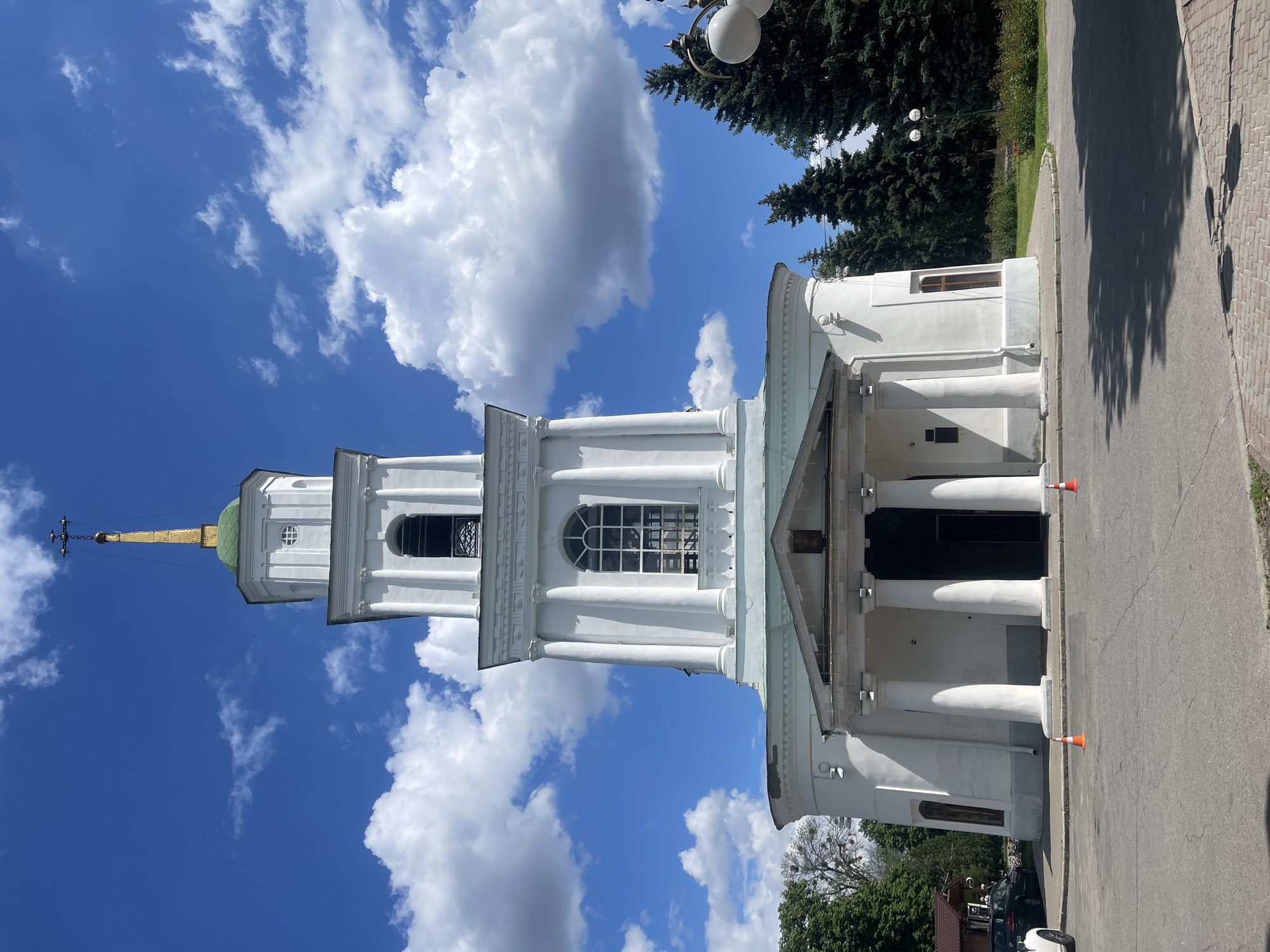 The image displays a tall, white church or cathedral under a bright blue sky filled with scattered white cumulus clouds. The main structure features a prominent bell tower with multiple tiers, topped by a light green dome and a golden spire culminating in a dark cross. The church facade exhibits classical architectural elements, including numerous white columns (likely Doric or Tuscan style) and large arched windows with gridded panes.
On the ground level, a paved road or path with reddish-brown paving stones runs alongside the building. Two orange and white traffic cones are visible near the church's entrance, suggesting a temporary demarcation or safety measure. To the right of the church, dense green conifer trees (possibly fir or spruce) line the area, with a decorative streetlight featuring two white globe lamps peeking out from among them. Parts of parked cars are visible in the lower right corner of the frame.
The photograph was taken on a clear, sunny day, indicated by the bright illumination and distinct shadows cast on the ground from the trees and building. No people are visible, and no specific activity is taking place. A horizontal band on the bell tower, beneath the largest arched window, contains some inscribed text, likely in Cyrillic script, but it is not clearly legible from this distance. The overall impression is one of a serene, historical architectural site on a pleasant day.