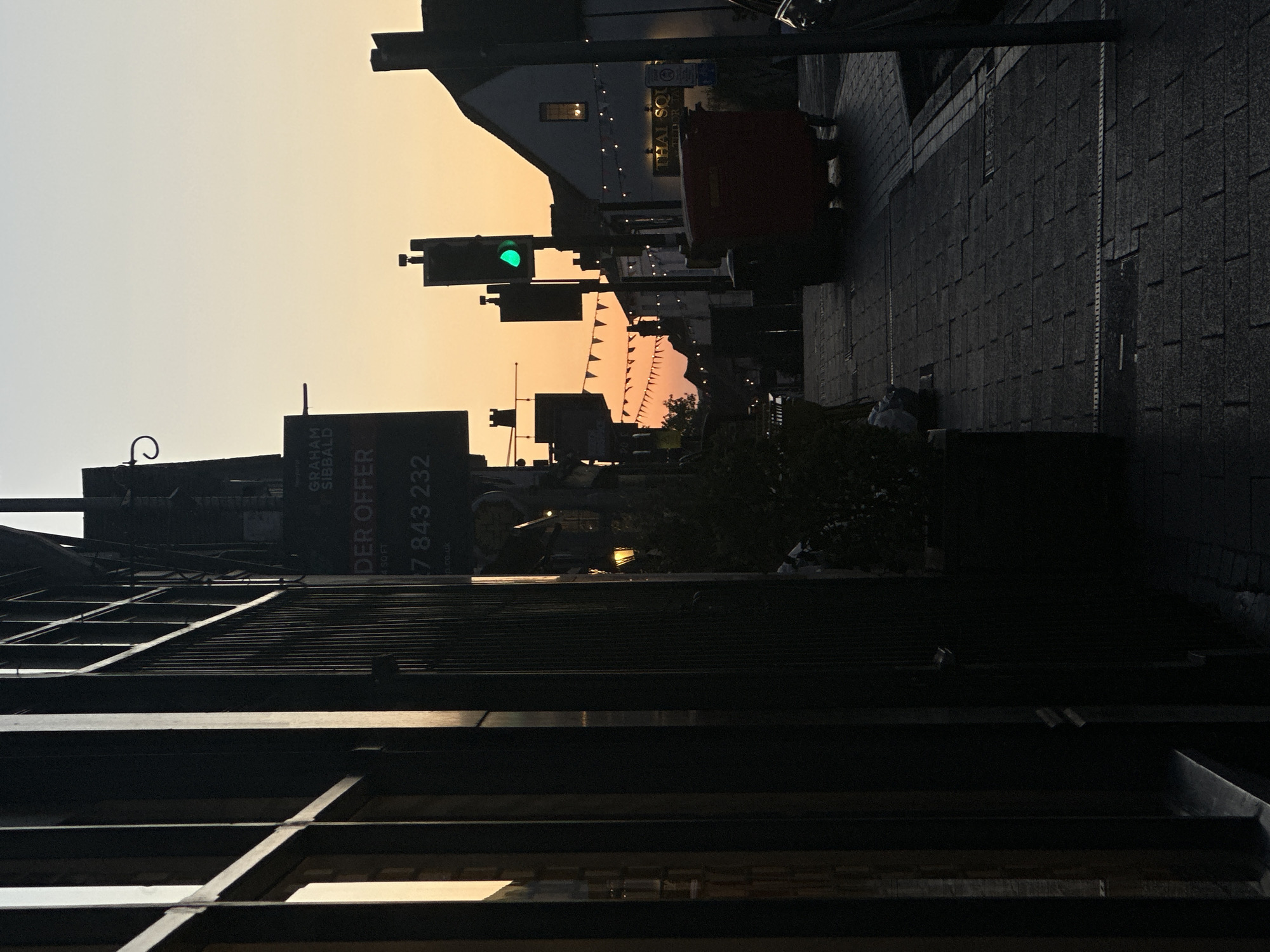 The image depicts a street scene in St Albans, United Kingdom, at dusk or dawn. The view is looking down a street lined with buildings. A traffic light is illuminated with a green light. Pennant flags are strung across the street. A sign for "Graham Sibbald" is visible on a building. The sky is a gradient of yellow and light grey. The street is paved with cobblestones.