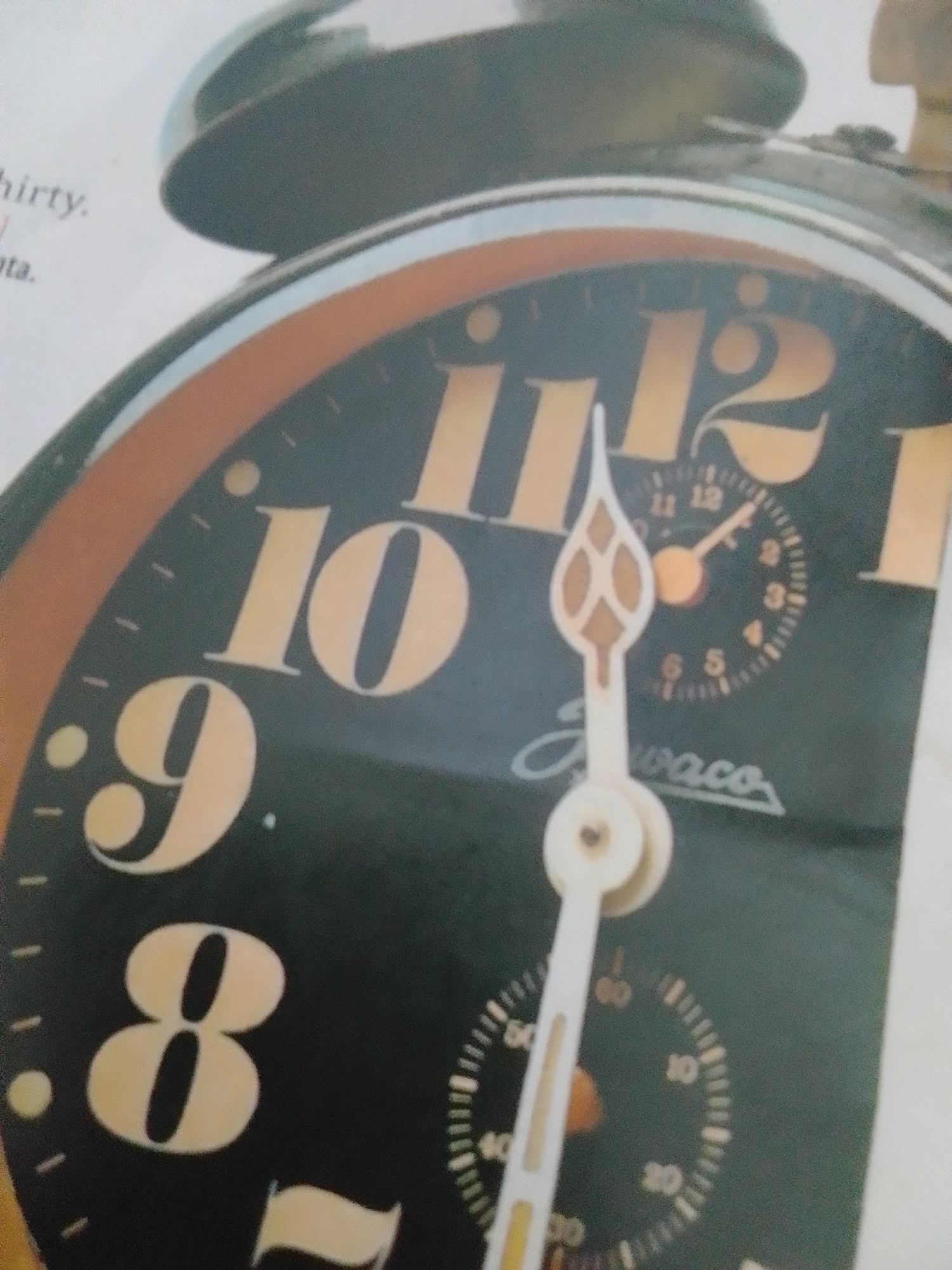 This is a close-up, angled photograph of a vintage alarm clock. The clock face is black with large, luminous yellow numerals marking the hours 8, 9, 10, and what appears to be 11 and 12. The hands of the clock are white, with the hour hand pointing close to the 9 and the minute hand pointing just past the 10. A smaller dial on the clock face shows what seems to be a stopwatch function with markings up to 60. The word "waco" is visible in a stylized script beneath the clock hands. The top of the alarm clock, with its metal bells, is also visible in the background. Text from a page is visible to the left of the clock, with partial words like "hirty." and "ata." suggesting it might be from a magazine or book. The lighting is bright, and there are no explicit environmental cues to suggest a specific time of day, weather, or location, other than the mention of La Tagua, Venezuela as a location context, which is not visually discernible in the image itself. The focus is entirely on the clock.