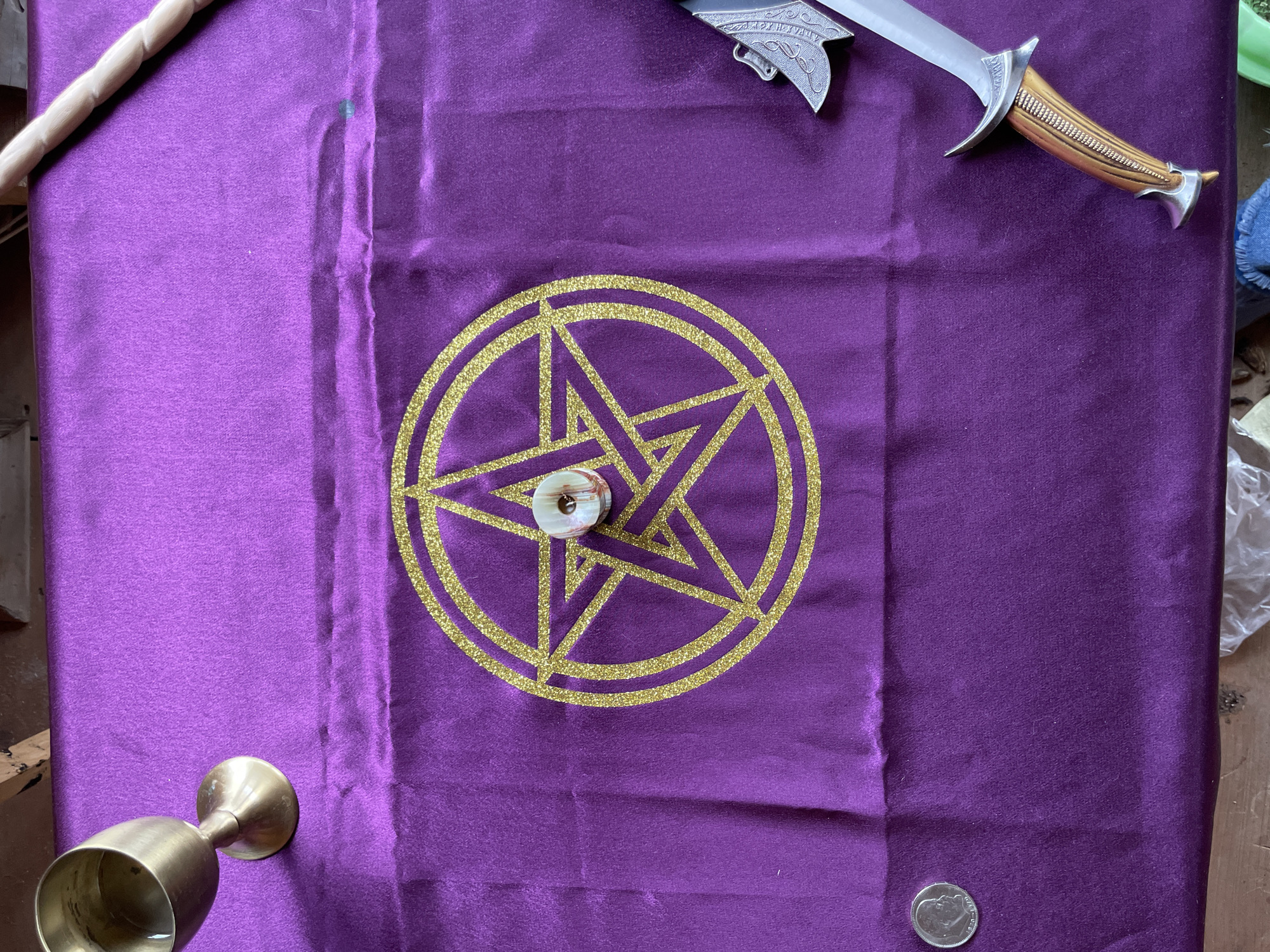 A purple cloth with a gold pentagram design in the center is laid out on a surface. A small, round, tan and brown object with a hole in the center rests on the pentagram. A wooden wand is in the upper left corner, and a dagger with a silver blade and carved wooden handle is in the upper right. A small gold chalice sits on the lower left, and a silver coin is in the lower right corner of the cloth. The scene appears to be set indoors. There is no visible information to indicate that this scene is in Zav'yalovo, Russia. There are no people in this image.