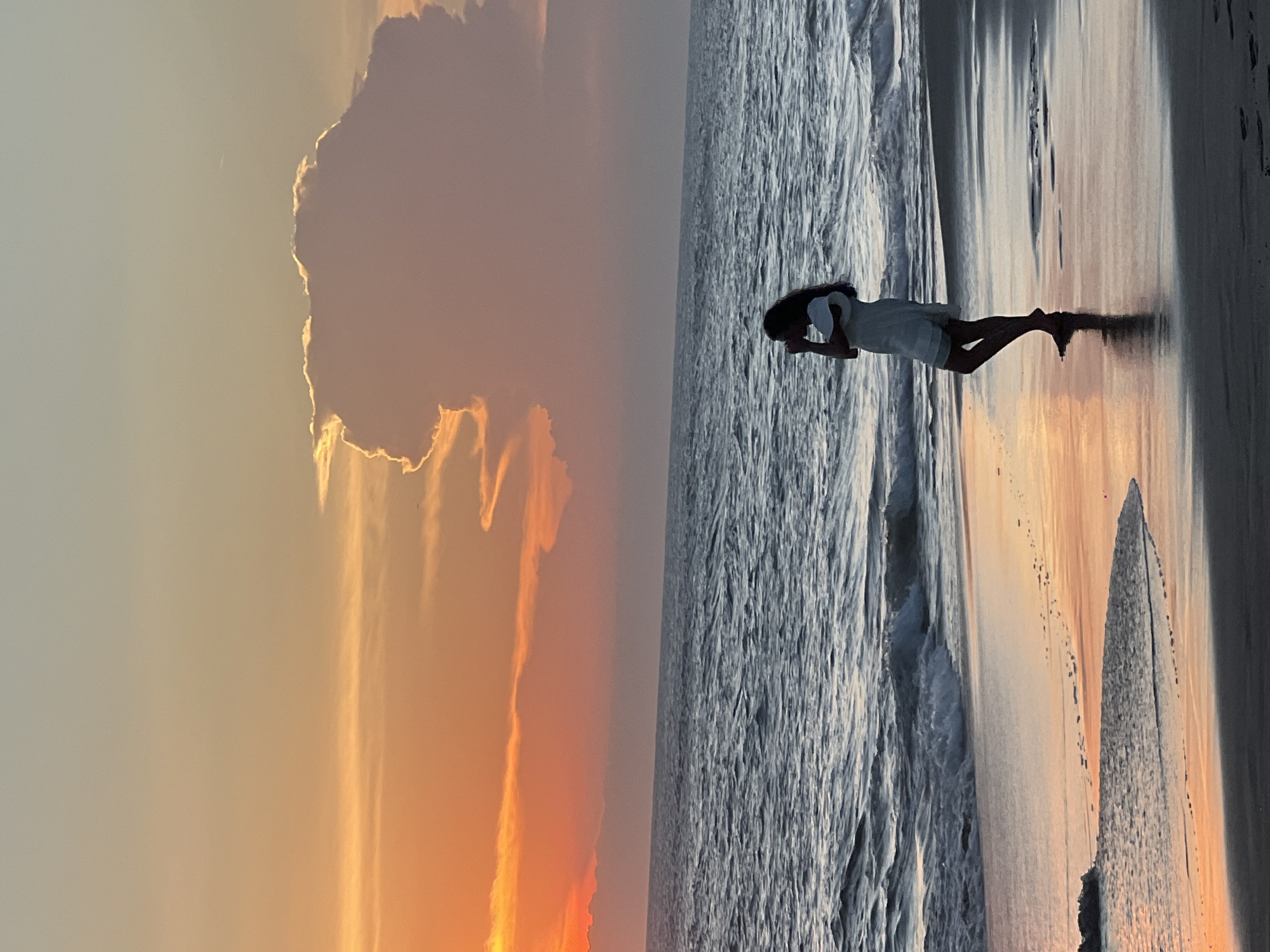 The image shows a beach scene at sunset in what is likely North Kuta, Indonesia. A person stands on the wet sand, facing towards the ocean. The sky is filled with orange and yellow hues from the setting sun, with cloud formations visible. The ocean waves are prominent, and there are footprints in the sand near the water's edge. The lighting suggests it is dusk or dawn.