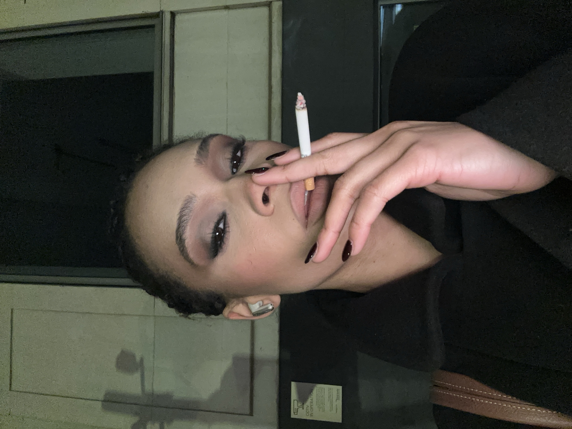 A woman is shown from the chest up, holding a lit cigarette to her lips with her right hand. She has dark hair pulled back, dark eye makeup, and dark nail polish. She appears to be wearing an earpiece in her left ear. She is wearing a dark-colored top or coat. In the background, there is a wall with vertical lines, and a sign that reads "CCTV in operation". A dark shape is behind the woman, possibly a building or other structure. It is impossible to determine if the location is in London, United Kingdom.