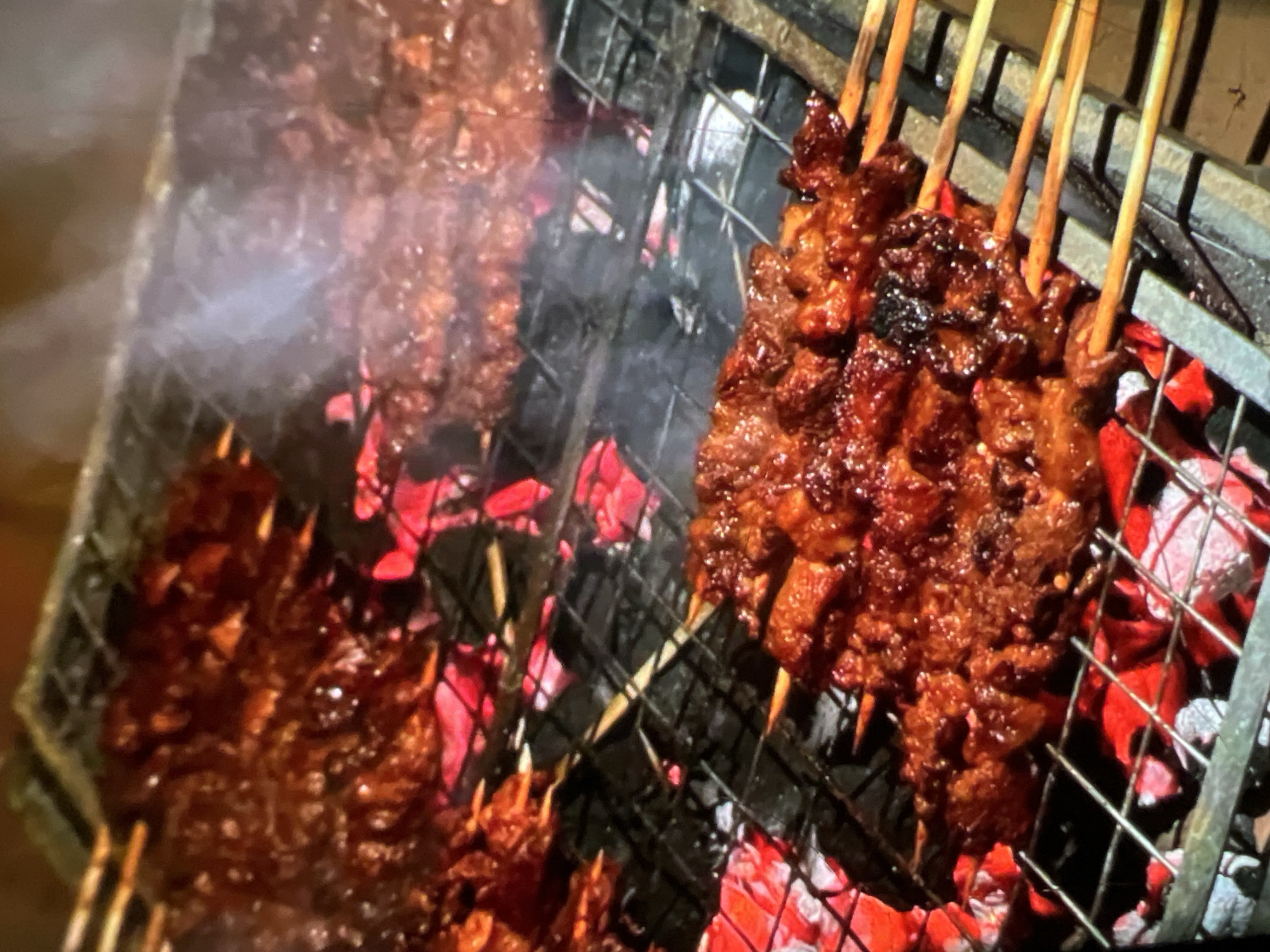 The image depicts a close-up view of meat skewers grilling over hot coals. The skewers, loaded with pieces of marinated meat, are arranged on a metal grill. Glowing red coals are visible beneath the grill, indicating high heat. Wisps of smoke rise from the cooking meat, suggesting it is actively being grilled. The scene appears to be outdoors, likely a street food vendor stall, given the rustic setup and the type of food preparation. The lighting suggests it could be nighttime or dusk, with artificial light illuminating the grilling process. While the image does not explicitly confirm a location in Lagos, Nigeria, the type of food and grilling method are common street food practices found in the city.