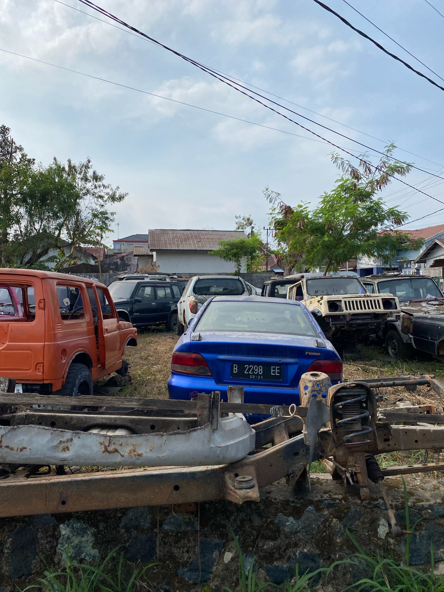 This is a vertical, eye-level, medium shot of a car junkyard in the city of Banjarbaru, Indonesia. The foreground is dominated by the chassis of a stripped-down vehicle. The chassis is rusted and sits atop a low stone wall. Behind the chassis are several vehicles in various states of disrepair. To the left, an orange, two-door Suzuki Jimny is missing its doors and hood. Further back, a black SUV, a white Jeep-like vehicle, and a blue sedan are visible. The blue sedan has a license plate that reads "B 2298 EE 02-21". In the background, houses with corrugated metal roofs and trees are visible under a cloudy sky. Overhead power lines stretch across the frame. The ground is covered in dry grass and dirt. There are no people visible in the image.